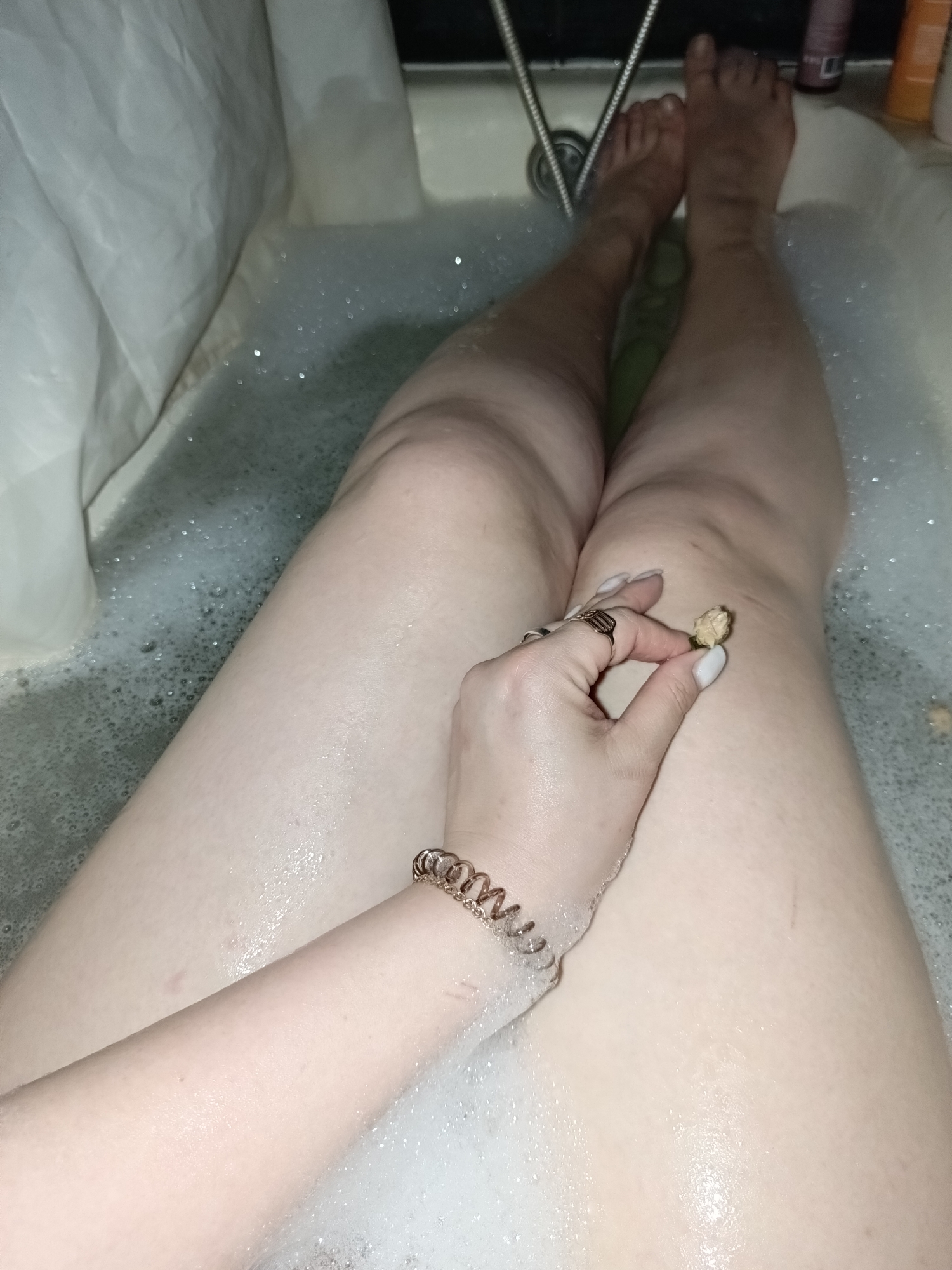 A person is captured from a first-person perspective, relaxing in a bubble bath. The central focus is on their lower body, specifically their legs and one arm, partially submerged in the foamy water of a light-colored bathtub. The person's skin appears fair, and light-colored nail polish is visible on their fingers and toes.
On the left side of the frame, a white shower curtain hangs. A metallic shower hose and head are visible near the feet at the far end of the tub. Several bath product bottles, including one purple and one orange, are arranged on the tub's edge.
The person's right hand, adorned with a ring on the middle finger and a spiral-shaped hair tie or bracelet on the wrist, gently holds a small, dried, light-colored flower bud on their knee. Another similar bud floats in the bubbly water nearby, suggesting a decorative or aromatic element to the bath. The water is filled with numerous white bubbles, reflecting ambient light.
The scene conveys a sense of personal relaxation and self-care in an indoor bathroom setting. The lighting is soft, contributing to a serene atmosphere. No text is visible in the image.