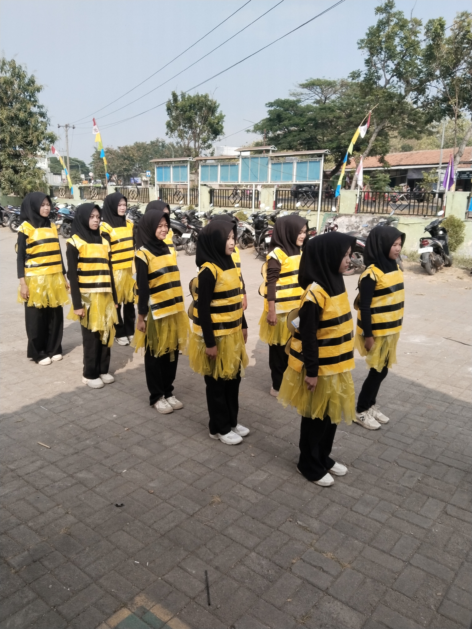 In Pakulaut, Indonesia, a group of eight young girls, dressed in black pants and black hijabs, stand in formation facing right. They are wearing bee costumes consisting of yellow vests with black horizontal stripes and fluffy yellow skirts. The background shows a street with parked motorcycles, a fence, trees, and a building. The ground is paved with rectangular bricks. The girls are posed as if for a photograph or performance.