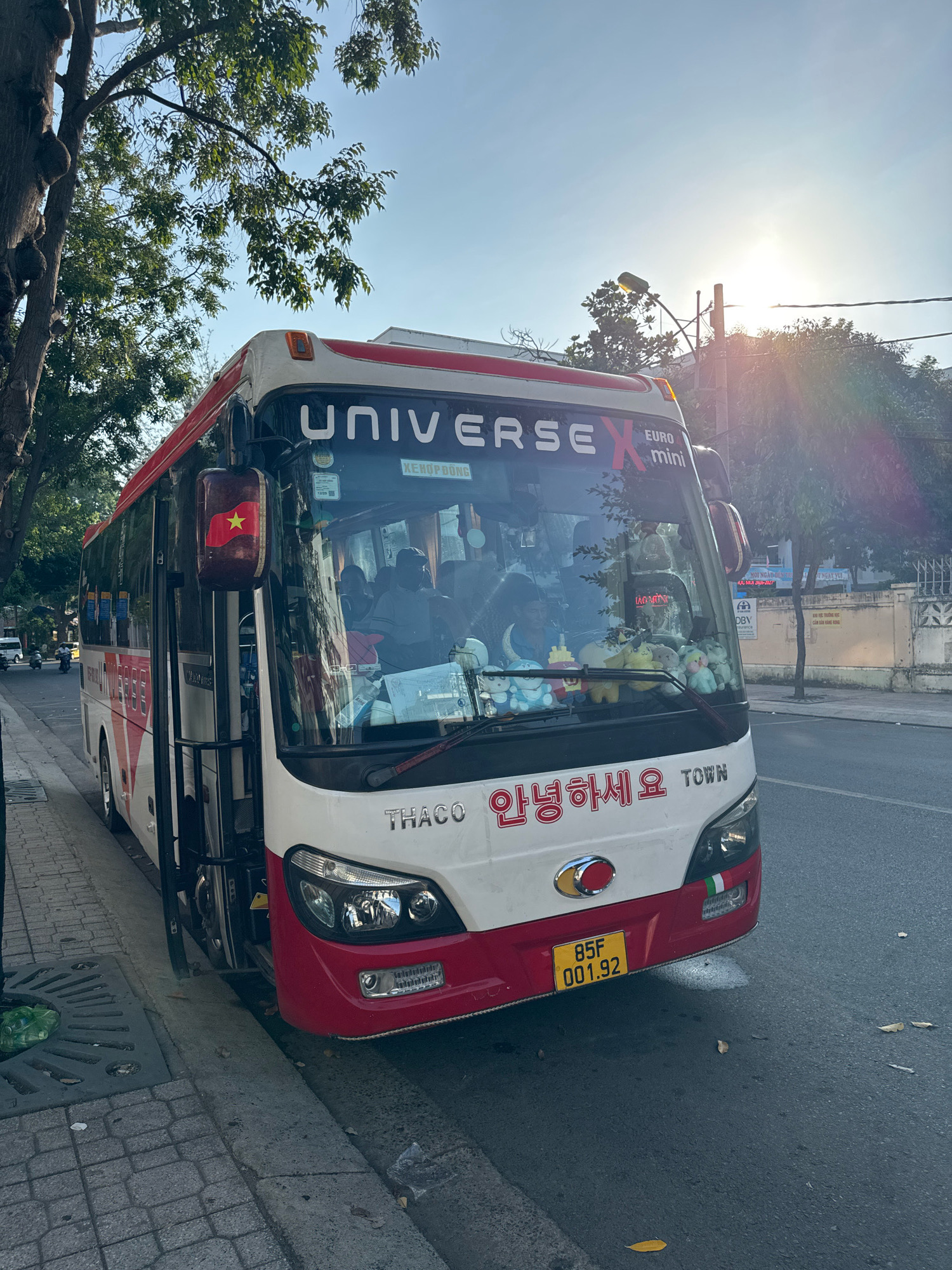 A white and red bus with "UNIVERSE X EURO mini" written on the windshield is parked on the side of a street in Nha Trang, Vietnam. The bus has a Vietnamese flag on its side mirror. The windshield is decorated with various stuffed animals. Korean text "안녕하세요" and "TOWN" are written on the front of the bus, along with the brand "THACO". The license plate reads "85F 001.92". The street is paved with asphalt, and there is a sidewalk with a concrete drainage grate on the left side of the image. Trees with green leaves are visible in the background. The sun is shining brightly in the sky, creating lens flare.