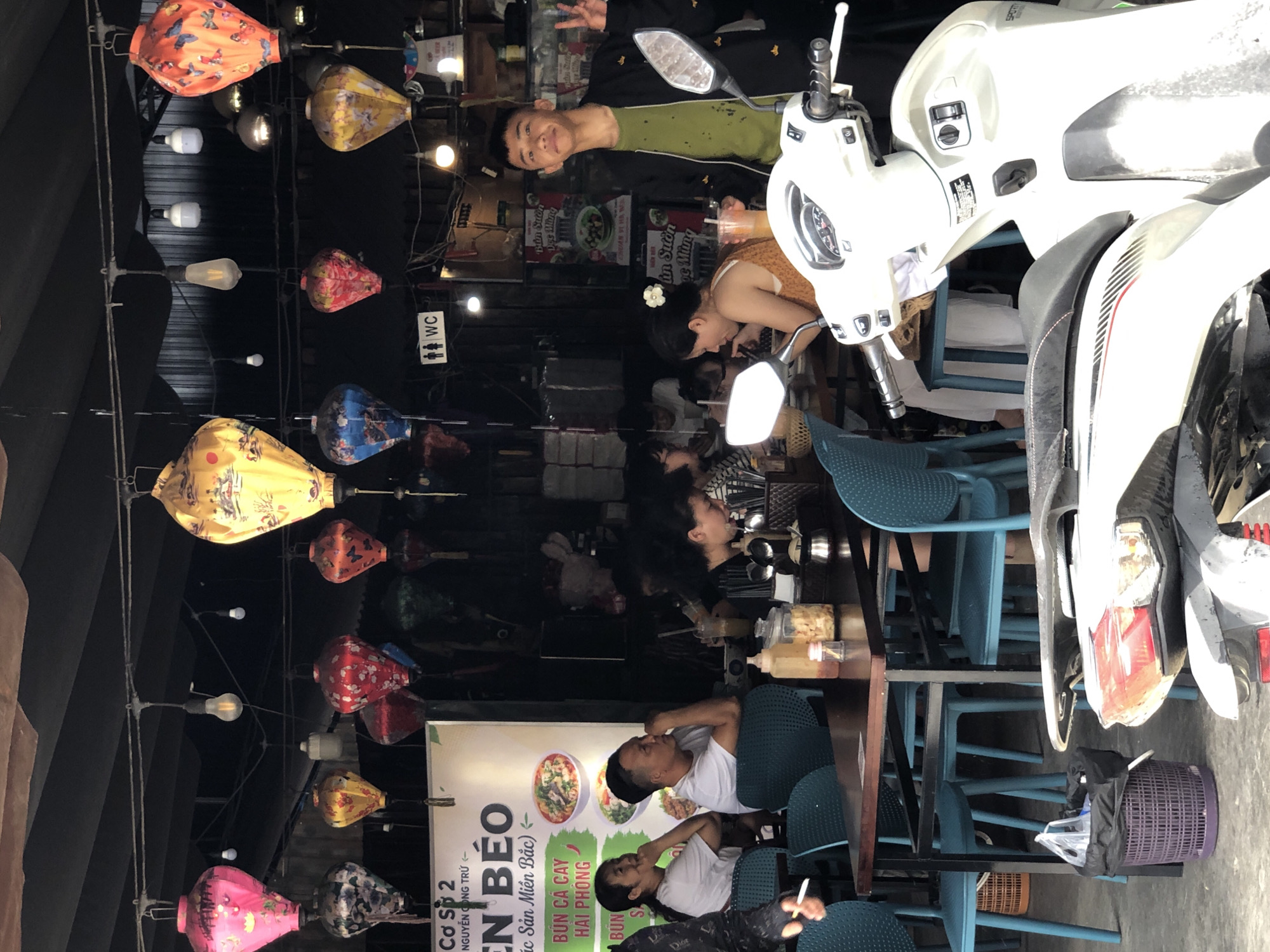 This is a street scene in Da Nang, Vietnam, featuring an outdoor eatery with customers dining and a young man posing near a white scooter. Lanterns are hanging overhead, and a sign on the wall advertises "BUN BO" (beef noodle soup). People are seated at tables, eating and drinking. The overall atmosphere suggests a casual dining environment in a bustling urban area.