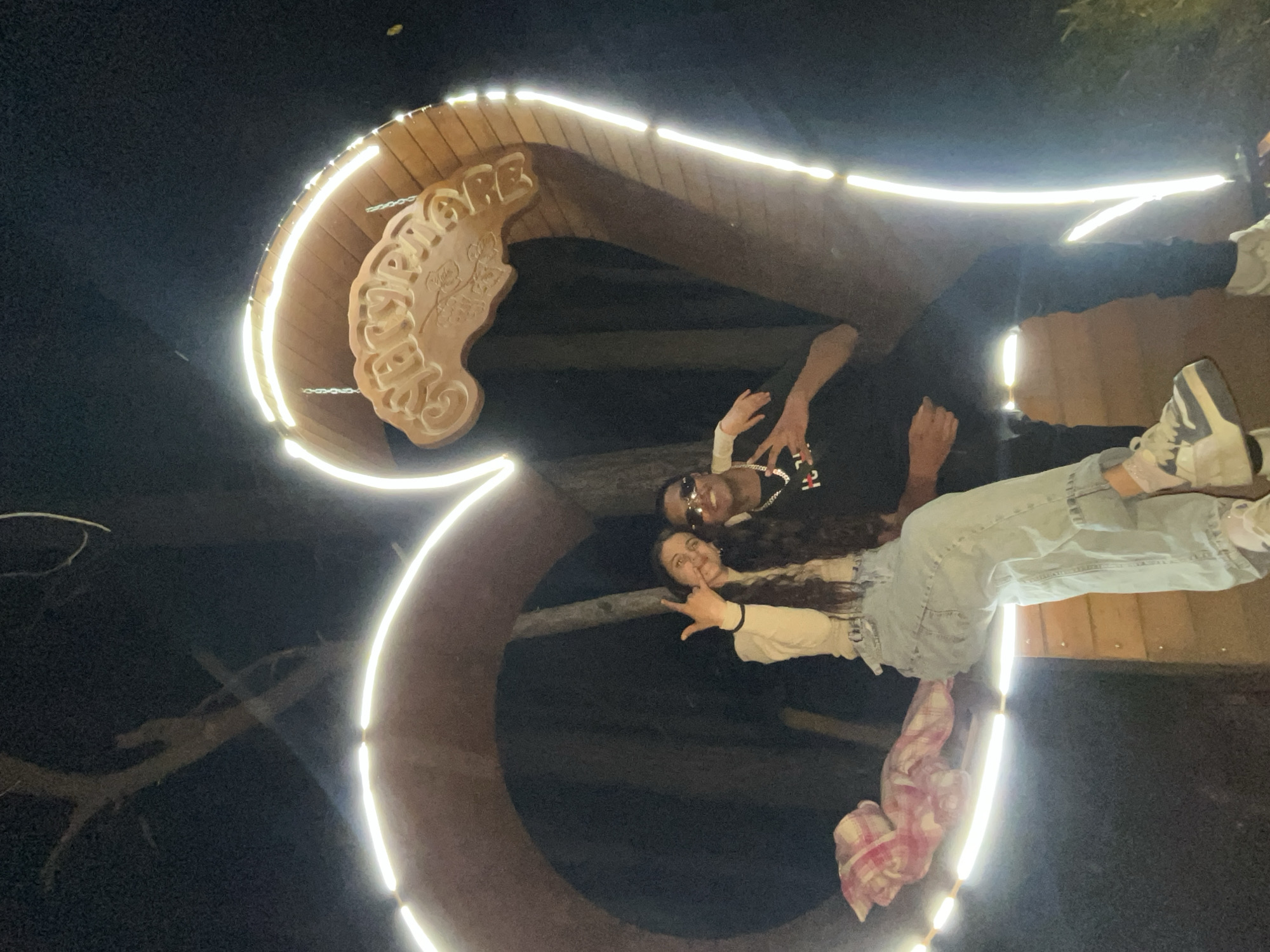 A nighttime shot of a couple posing in front of the entrance sign of Sungurlare, Bulgaria. The entrance is a wooden arch with illuminated LED lights around the frame, with the town's name in a wood-carved sign. The woman is on the right, wearing jeans and making the "rock on" hand gesture. The man is above her, wearing sunglasses and a T-shirt. They appear to be standing inside the arch. The background is dark.