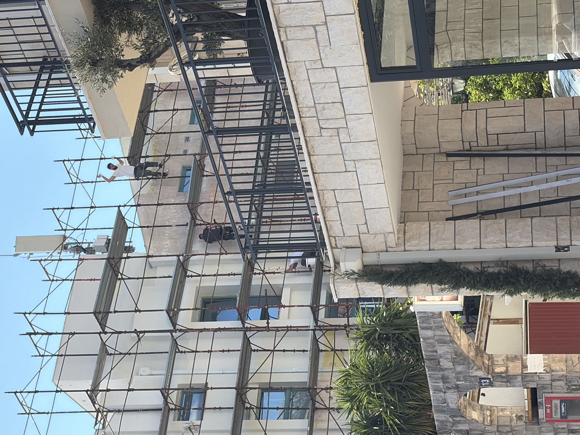 The scene depicts the exterior of a building in Budva, Montenegro. The building is undergoing renovations, indicated by the extensive scaffolding erected along its facade. Two individuals are visible on the scaffolding, apparently engaged in construction or maintenance work. The building features a light-colored, possibly stone, facade. There's a balcony with a dark railing extending from the building. A small tree or shrub is planted on the balcony. Near the base of the building, there are more trees and a portion of another building.