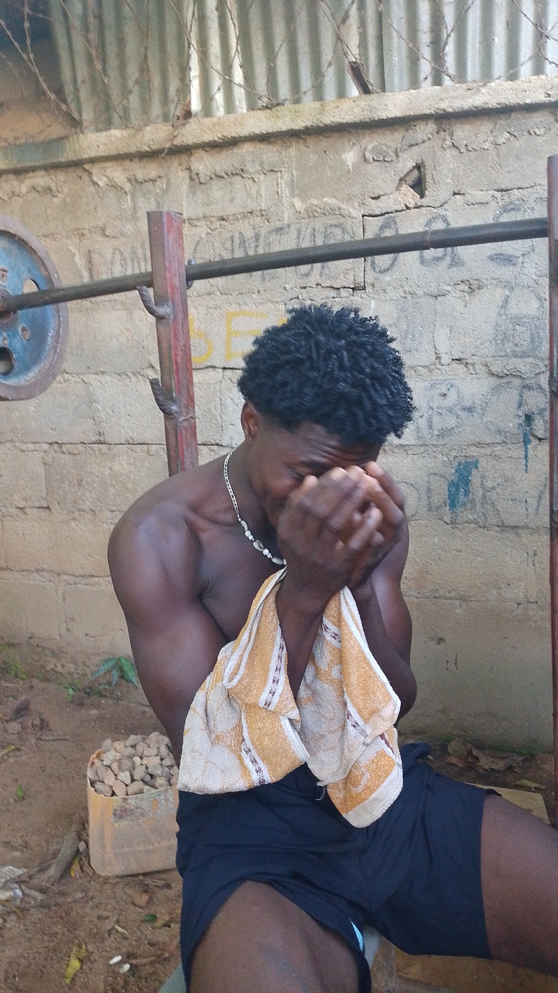 A young Black male is sitting outdoors, shirtless, with a towel draped over his lap. He is wearing black shorts and a silver necklace with a shell-like pendant. His hands are covering his face, and he appears to be crying or distressed. The background consists of a rough, concrete block wall, a metal weightlifting bar with a blue weight plate, and a rusted corrugated metal fence above the wall. A small, orange plastic container filled with stones or rocks is on the ground to the left of the man. The ground is dirt with some scattered leaves. The overall impression is one of a gritty, outdoor workout space, possibly in a less developed area.