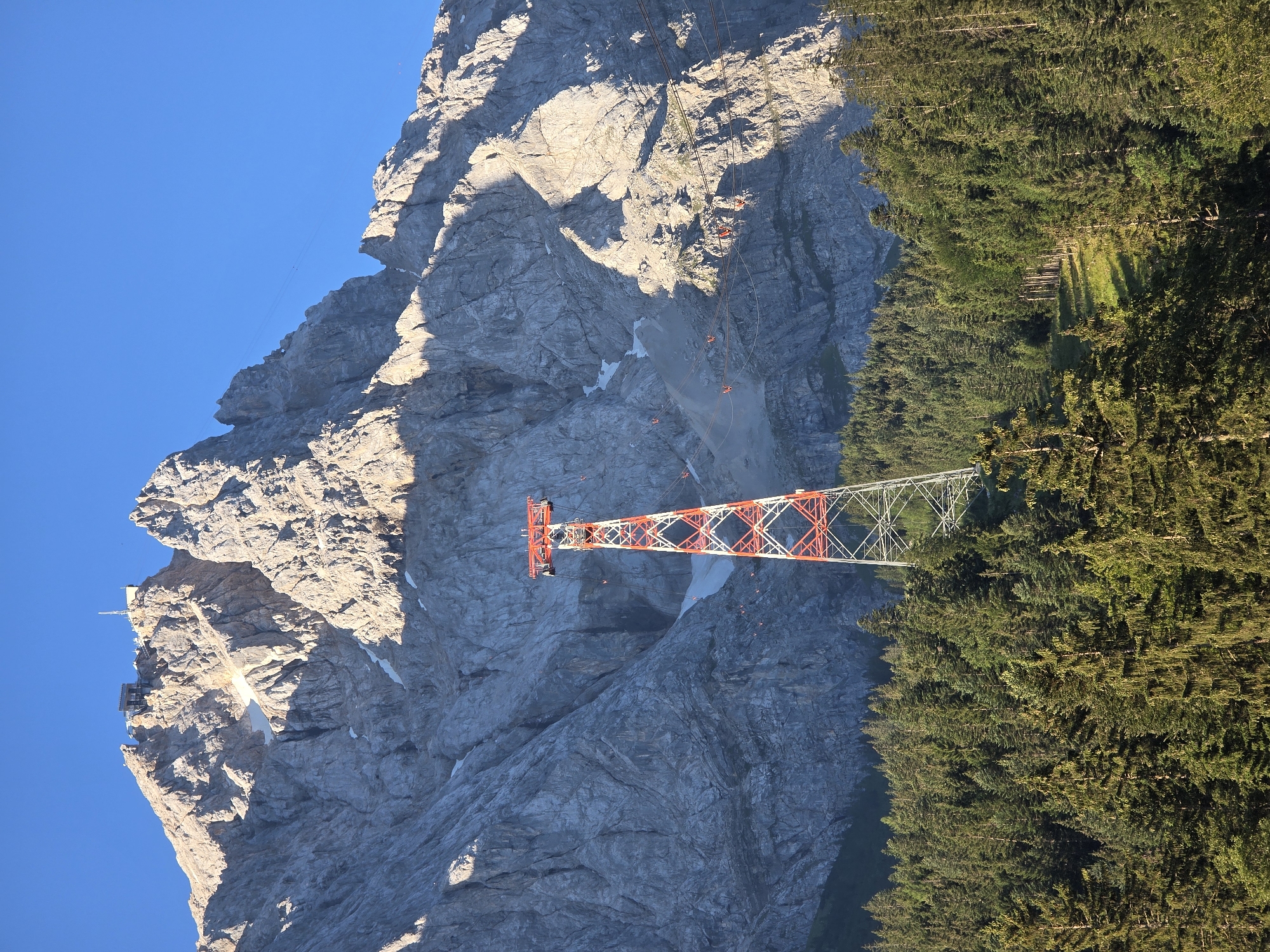 The image depicts the Zugspitze, Germany's highest mountain, with a cable car tower in the foreground. The tower is a lattice structure painted red and white. A lush green forest of pine trees covers the lower slopes of the mountain. The upper reaches of the mountain are rocky and barren, with patches of snow visible. A building and antenna are situated on the summit. Several red cable car cabins are visible ascending the mountain. The sky is a clear, vibrant blue. This scene is located in the municipality of Grainau, Germany, and captures the natural beauty and infrastructure associated with the Zugspitze massif.