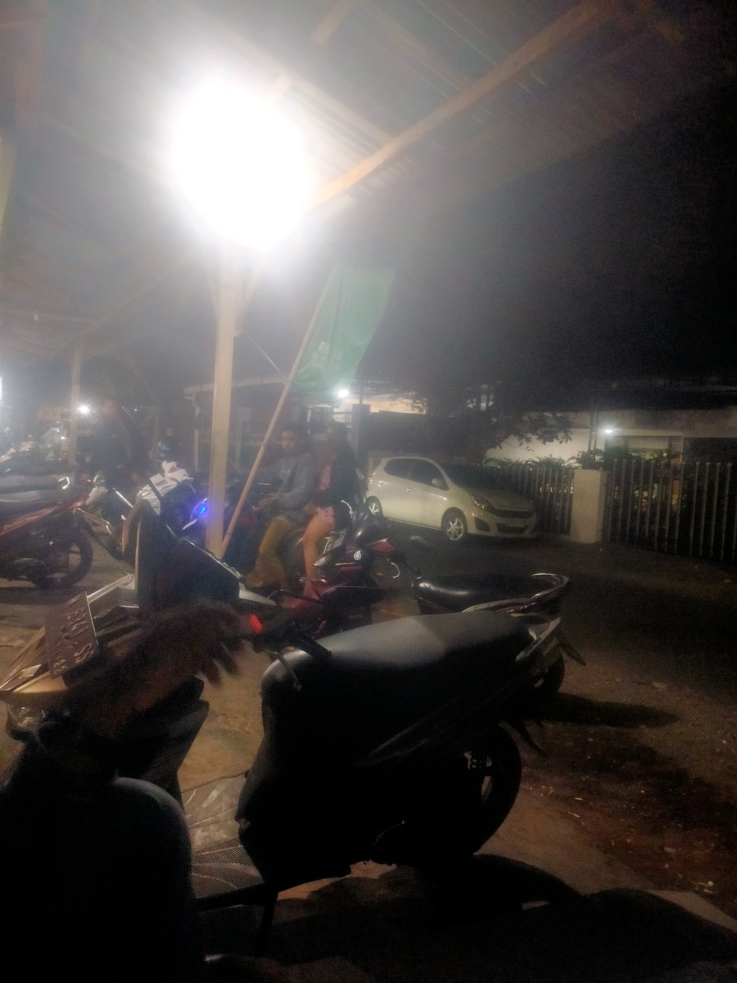 The image depicts a dimly lit outdoor scene, likely in Lumajang, Indonesia, at night. A bright overhead light illuminates a covered area where several individuals are gathered around motorcycles. In the foreground, the silhouette of a person and a motorcycle are prominent. Further back, other motorcycles are parked, and two individuals are seated on one. A white car is visible in the background, parked in front of a fence. The overall atmosphere is casual, suggesting a gathering or waiting area.
