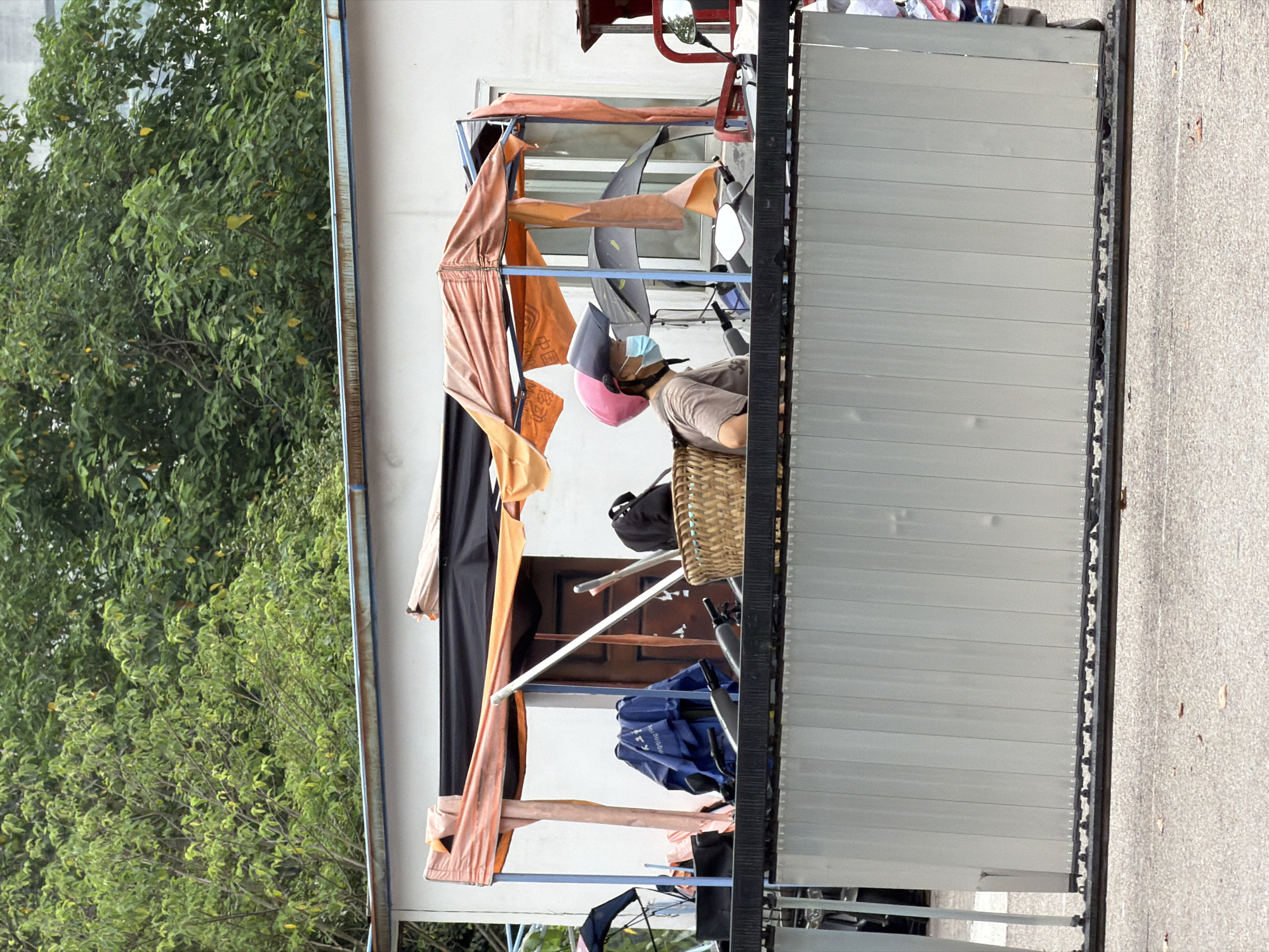 A side profile of a person wearing a pink helmet and a light blue face mask is visible in the center of the frame. The person is seated on a scooter, partially obscured by a straw basket and an orange canopy. The person's torso is clad in a brown t-shirt. Behind the person, a wooden door is visible within a white building. A blue garment is draped over a scooter's handlebars to the left of the person. The scene appears to be an outdoor market stall, indicated by the temporary canopy structure and the display of goods. In the background, lush green trees provide a natural backdrop. A grey, corrugated metal barrier runs across the foreground, separating the market area from a paved surface. The overall environment suggests an urban setting, possibly in Jiangjin, China, given the context provided.