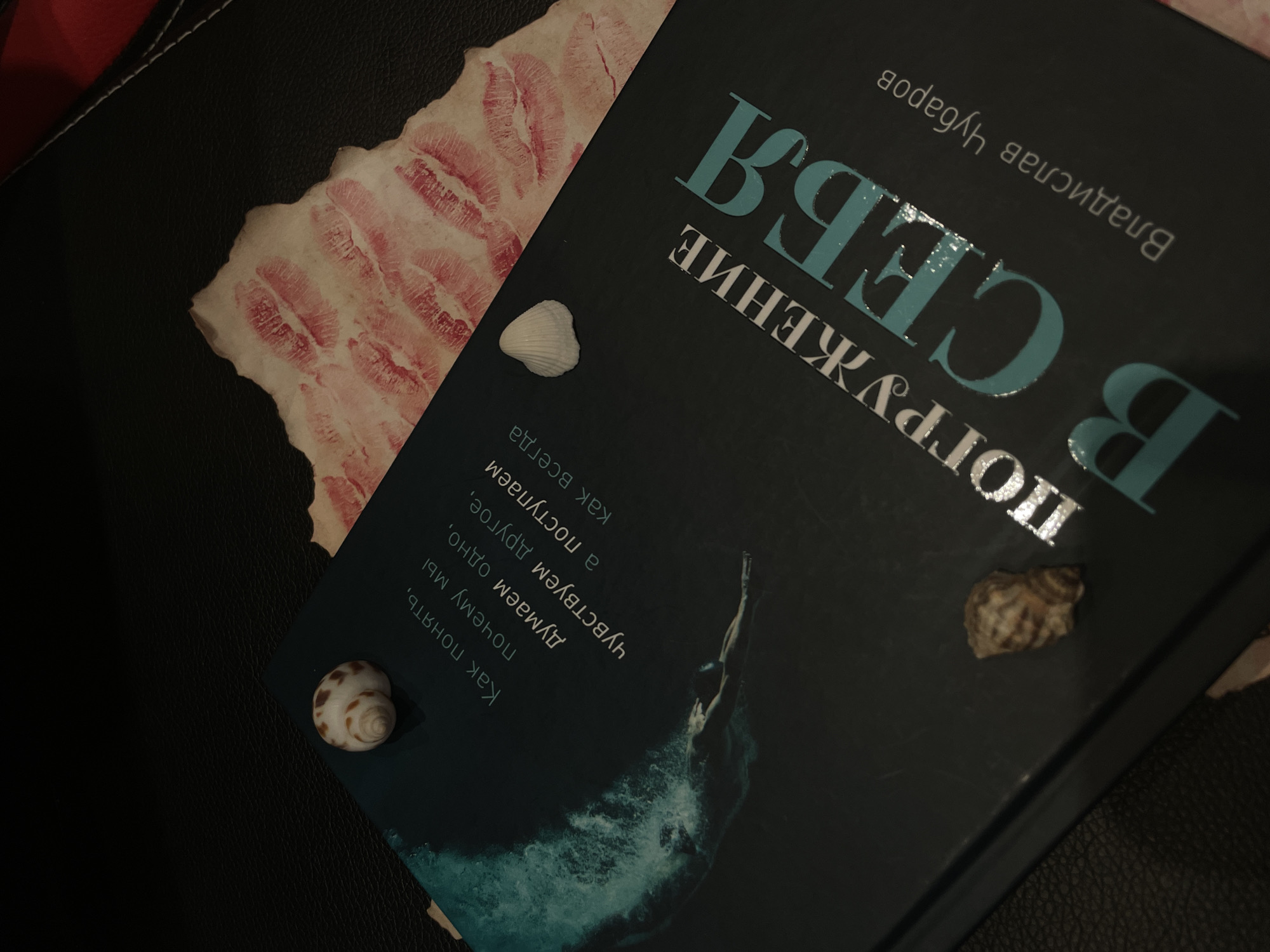 The image is a close-up, angled overhead shot of a book titled "ПОГРУЖЕНИЕ В СЕБЯ" (Immersion in Oneself) by Владислав Чубаров (Vladislav Chubarov). The book is dark blue and features a graphic of a person diving into water on the left side. Text on the book reads: "Как понять, почему мы думаем одно, чувствуем другое, а поступаем как всегда". Two shells are placed on the book, one on the upper left and another on the upper right. Behind the book, a torn piece of paper with numerous red lipstick kiss marks is visible. The background is a dark, textured surface, possibly leather. The scene is dimly lit, with the light source seemingly from the top right, casting subtle shadows. There are no people present.