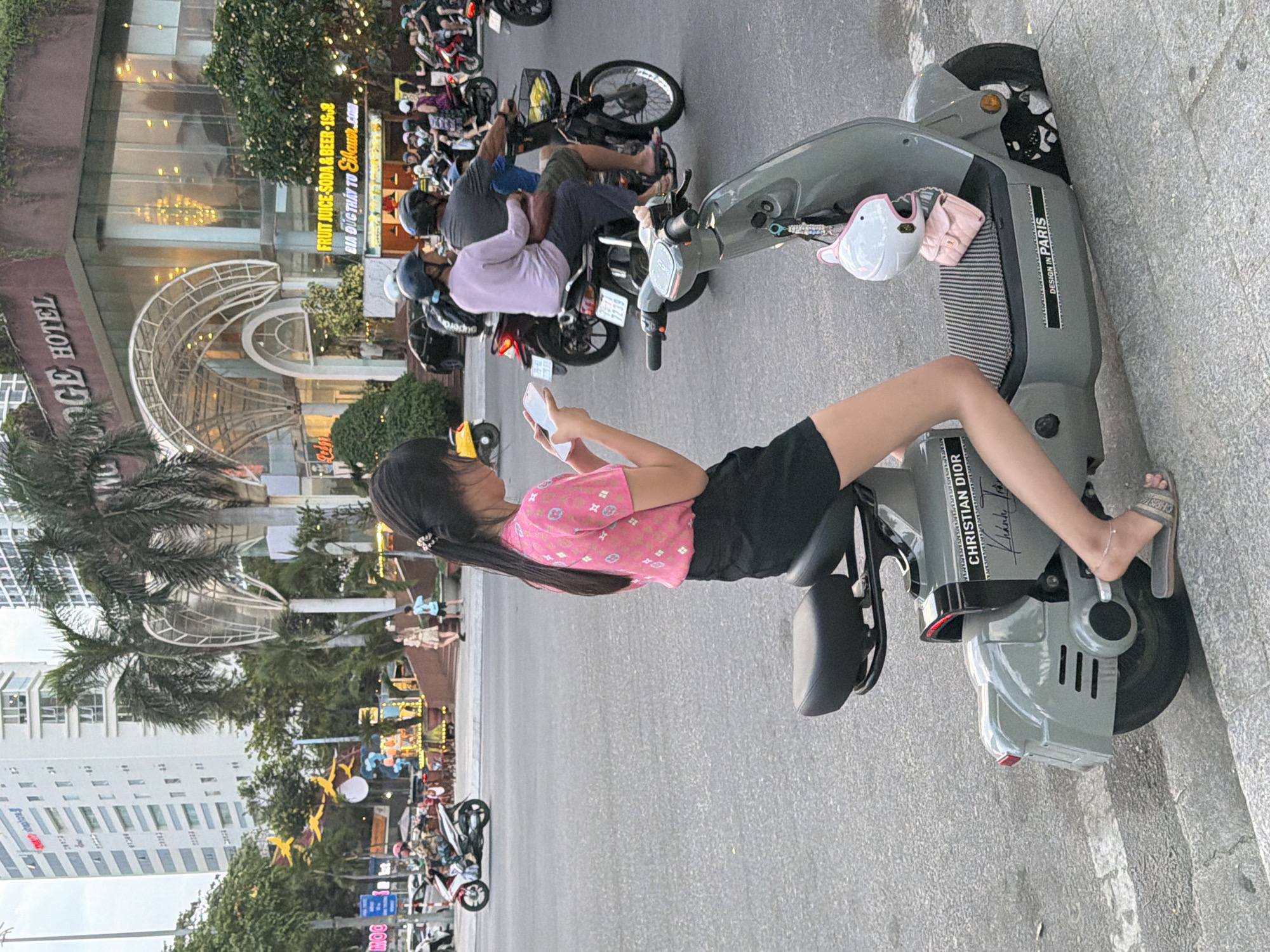 A young woman with dark hair, wearing a pink patterned shirt and black shorts, sits on a gray electric scooter in an urban street in Nha Trang Ward, Vietnam. She is looking down at her white smartphone. The scooter has a black seat, a white helmet with pink trim, and a pink purse hanging from its handlebars. The scooter also has "CHRISTIAN DIOR" and "DESIGN IN PARIS" stickers on its body. In the background, a building with the words "KING LODGE HOTEL" is visible, along with palm trees and other vehicles, including motorcycles. The street appears to be paved with asphalt and there are sidewalks on either side. The lighting suggests it is either early morning or late afternoon.