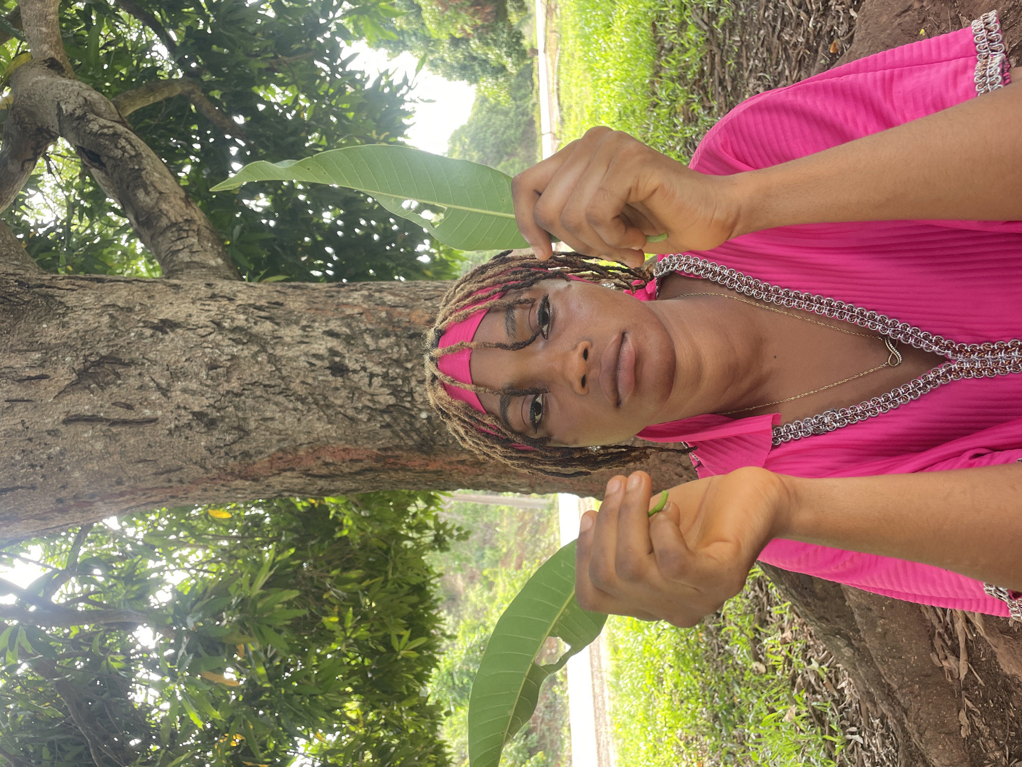 The image, viewed in portrait orientation, depicts a dark-skinned woman in an outdoor, natural setting, looking directly at the camera with a calm expression. She wears her brown dreadlocks pulled back with a bright pink headscarf or bandana, matching her vibrant pink, long-sleeved garment adorned with elaborate silver-colored trim along the neckline and cuffs. A gold-colored necklace with a heart-shaped pendant is visible around her neck. She holds two large, vivid green leaves, one in each hand, seemingly presenting or displaying them; these leaves strongly resemble mango leaves.
The background features a prominent tree trunk with rough, grey bark in the left foreground, which appears to be a mango tree. Its leafy branches extend upwards and across the frame, forming a dense canopy of green foliage through which bright, dappled sunlight filters. This indicates a sunny daytime, likely morning or afternoon. The right side of the background showcases more abundant green vegetation, including grass and undergrowth, suggesting a verdant, tropical environment consistent with the provided location context of Ogbe, Nigeria. There is no visible text in the image.