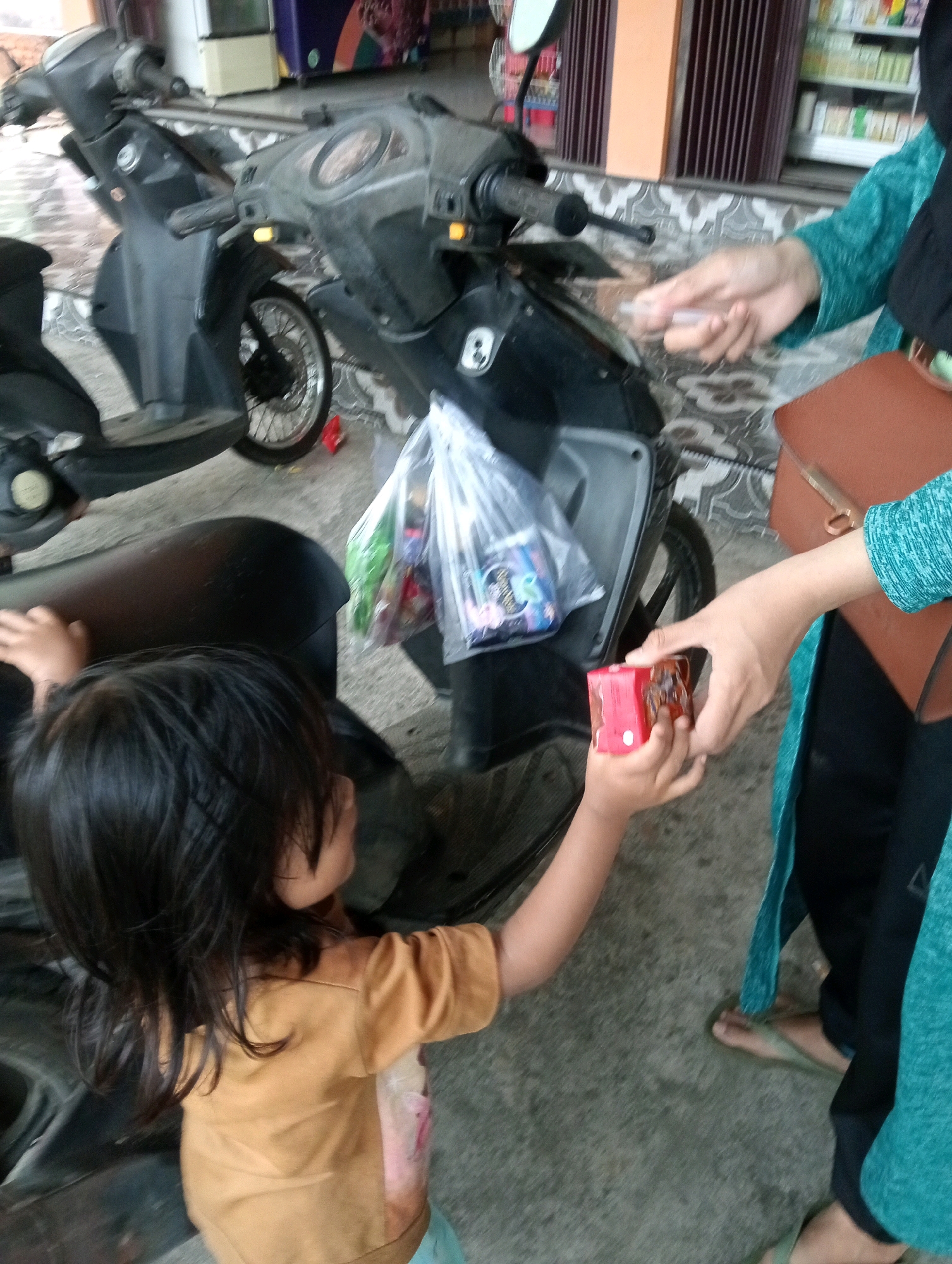 In front of a store in Campago, Indonesia, a person in a teal hijab and clothing extends a small red box to a child with long dark hair. The child, wearing a tan shirt, reaches out to take the box. A motorcycle is parked behind the individuals, and a plastic bag filled with items is visible on its seat. The ground is paved with gray tiles.