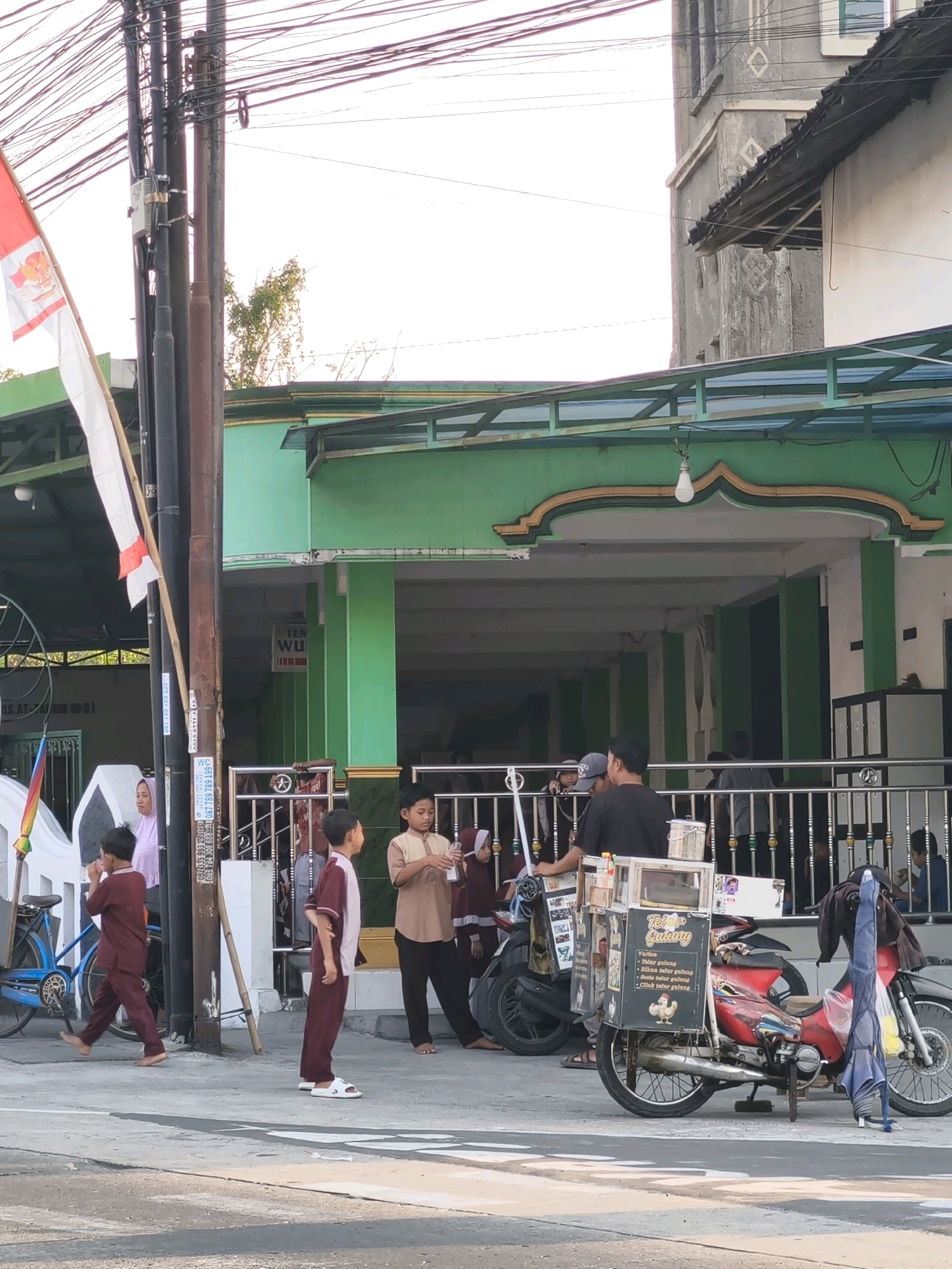 The image depicts a street scene in Desa Dibal, Indonesia, near a green building that appears to be a mosque. In the foreground, a red motorcycle with a food cart attached is parked. The food cart has signage that includes the text "Telur Gulung." Several people are present, including two young boys in maroon tunics and pants standing on the sidewalk, and a woman in a pink hijab standing near a bicycle. Another young boy in a maroon outfit is walking past the motorcycle. In the background, people are seen entering and exiting the green building. Overhead, a complex network of electrical wires is visible against a bright sky. A portion of a grey building with decorative diamond patterns is also visible on the right side of the image.