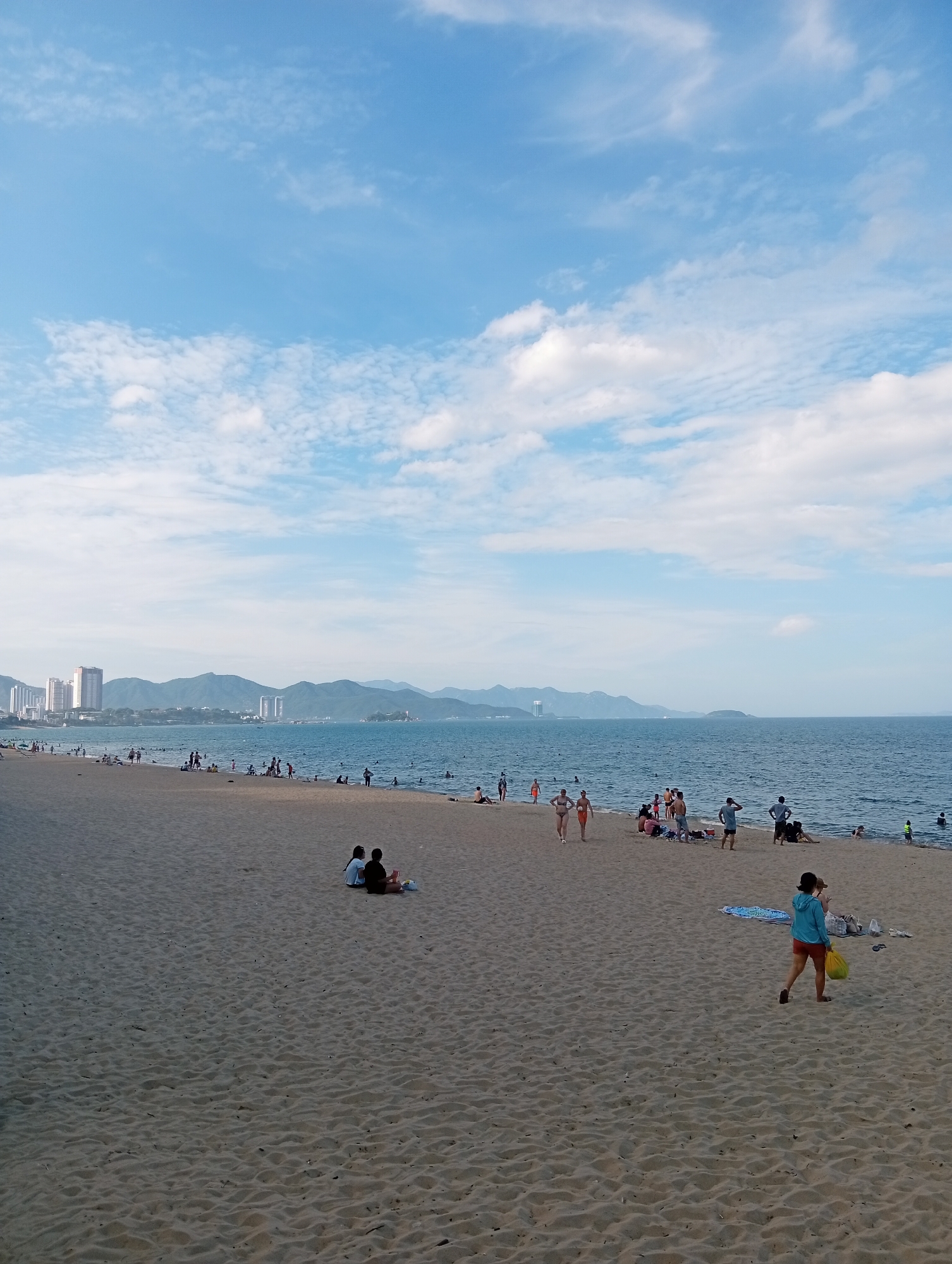 This media file depicts a crowded beach scene in Nha Trang, Vietnam. The time of day appears to be late afternoon, as indicated by the soft, diffused light and the shadows cast on the sand. The weather is pleasant, with a clear blue sky dotted with scattered white clouds, and the sea is calm and inviting.
The foreground is dominated by a wide expanse of golden sand, marked with the footprints and impressions of many visitors. In the middle ground, numerous people are engaged in various beach activities. Some are wading in the shallow water, while others are sunbathing or relaxing on towels and beach blankets. Several groups are gathered, chatting and enjoying the seaside atmosphere. Further back, a woman in a blue hoodie and orange shorts walks across the sand carrying a yellow bag. In the distance, the city skyline of Nha Trang rises against a backdrop of lush green mountains, with several tall buildings and structures visible. The ocean stretches out to the horizon, merging with the sky in a hazy blue expanse.
The overall mood is one of leisure and enjoyment, with people of all ages participating in a quintessential beach day. The scene captures the vibrant energy of a popular tourist destination, offering a glimpse into the daily life and recreational pursuits of the region. No visible text is present in the image.