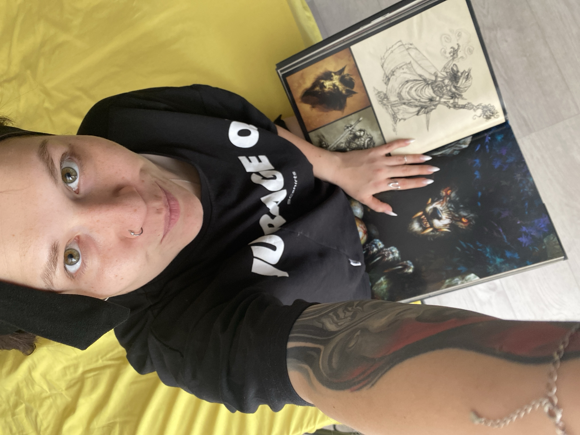 A selfie-style image captures a person lying down and looking directly at the camera with a calm, slightly smiling expression. The individual has light skin, green/brown eyes, freckles, and a silver nose ring. They are wearing a black short-sleeved t-shirt. Visible white text on the shirt reads "RAGE" with the beginning letters obscured, and below it, smaller text indicates "SC-SHIRTS". A prominent tattoo featuring dark and red swirling patterns covers a significant portion of their right arm, complemented by a thin silver chain bracelet on the wrist.
Their left hand, adorned with two silver rings and long white-tipped fingernails, rests on a large, open artbook. The book displays various illustrations: one page features a detailed pencil sketch of a flying or floating figure emitting smoke. Another page shows smaller images, including a creature's head in brown tones and a partially visible warrior figure. The book's open cover features a striking, dark illustration of a creature, possibly a wolf or monster, with glowing eyes amidst blue and purple foliage.
The person is positioned on a bright yellow surface, likely a blanket or bed sheet, with a light-colored floor visible beside the artbook. The scene depicts a relaxed indoor setting. While the user specifies the location context as Chaikovsky, Russia, this is not visually apparent in the image itself.
