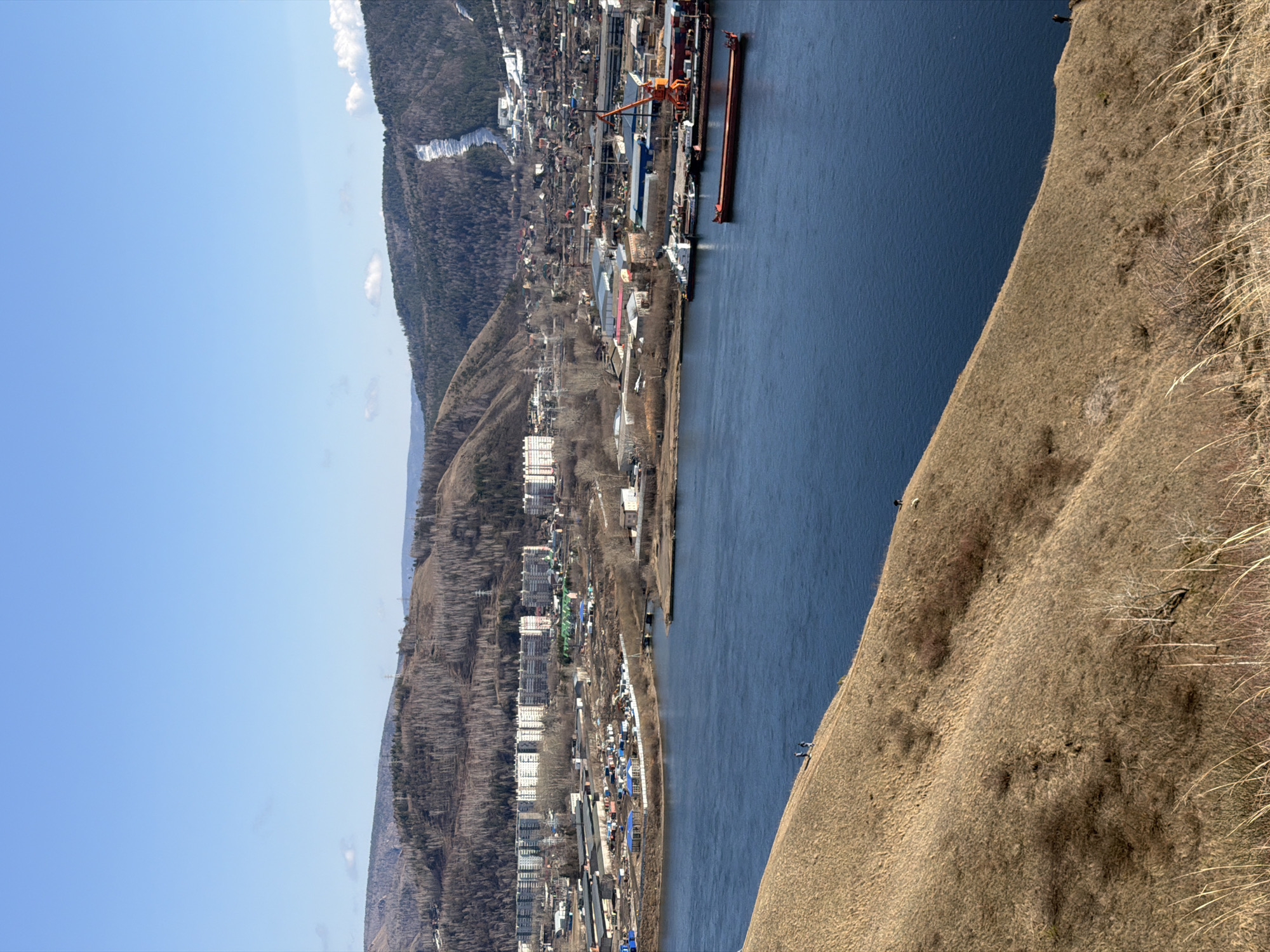 The image shows a view of Krasnoyarsk, Russia, from a high vantage point, potentially a hillside or mountain. In the foreground, there is a steep slope covered in dry grass. A large body of water (the Yenisey River) separates the slope from a town and industrial area on the opposite shore. The town consists of buildings, trees, and roads, with some taller apartment blocks visible. An industrial site features a loading crane and docked barges. A forested hill is visible behind the town and industrial zone. The sky is clear and blue.