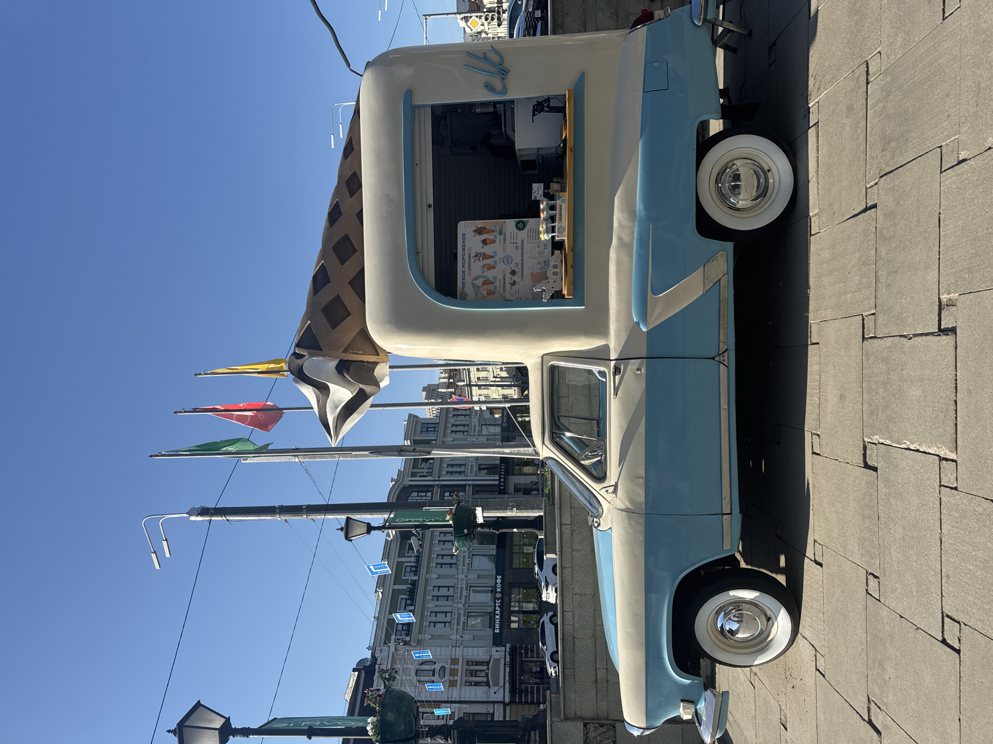 The image shows a vintage ice cream truck, painted in light blue and white, parked on a paved area that appears to be a pedestrian zone or sidewalk. The truck has a prominent ice cream cone replica mounted on its roof. The front of the truck faces the left.
Behind the truck, buildings line the street, indicating an urban environment. Street lamps and overhead wires are also visible. Some signage on the buildings is in Russian. The visible signage and architectural style suggest that the image was taken in Kazan, Russia.
The sky above is clear blue, suggesting a sunny day. No people are directly visible in the immediate foreground or around the truck. There is an ice cream cone menu visible inside the truck. Several flags are hung in the city behind the truck.