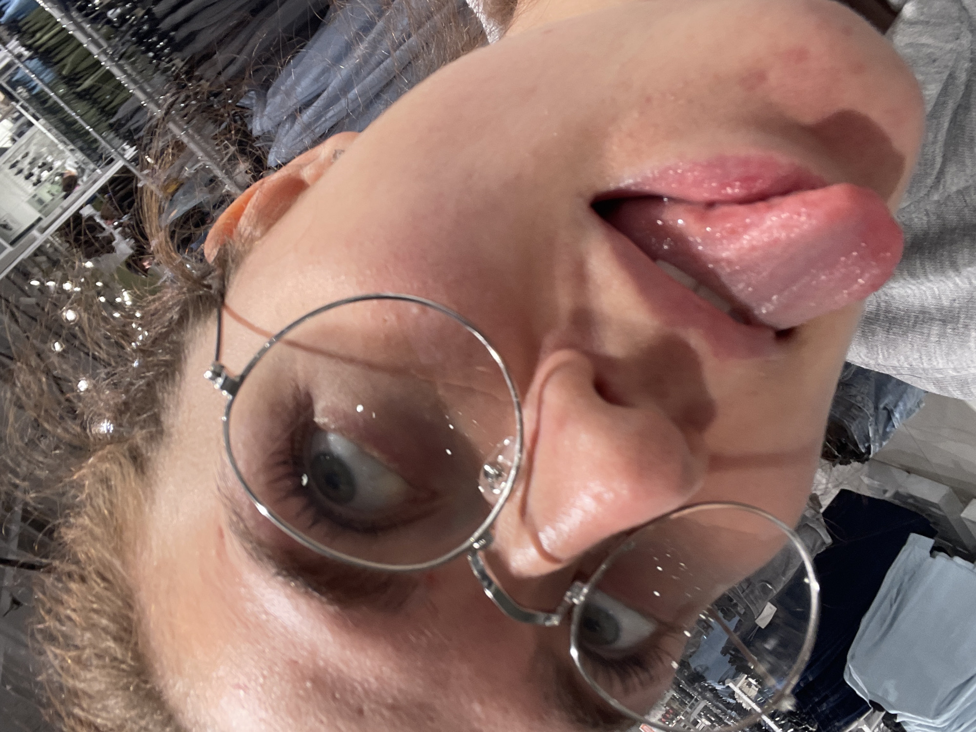The image presents a close-up, rotated perspective of a person's face. The individual has fair skin, wearing round, wire-rimmed glasses. Their tongue is extended. The environment appears to be a clothing store, evident from the reflected rows of clothing racks in the lenses of the glasses. Based on the location prompt, this clothing store is in Kommunarka, Russia.
