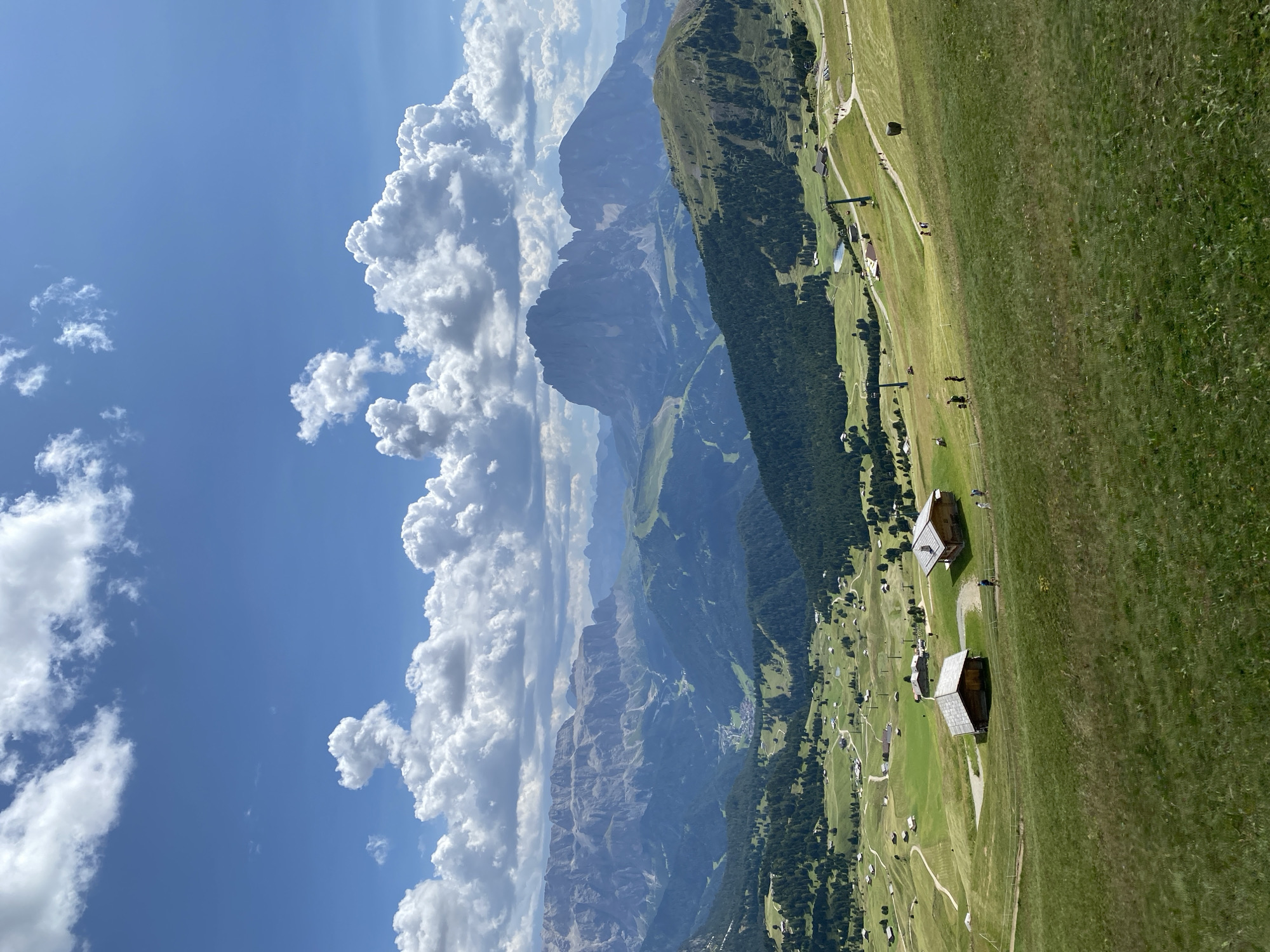This is an aerial landscape photograph taken from a high vantage point, looking down into a vast mountain valley under a blue sky with scattered white cumulus clouds. The foreground is a lush, green grassy slope dotted with wildflowers. In the middle ground, several rustic wooden chalets with grey roofs are nestled into the hillside, with a few people walking on paths between them. Further down the slope, a ski lift with several towers is visible, along with more chalets and a small lake. The valley floor is covered in dense green pine forest, with a small village visible in the distance. The background is dominated by imposing, rugged grey mountain ranges. The scene suggests a summer day in a mountainous region, likely the Dolomites in Italy, known for its dramatic peaks and green valleys.