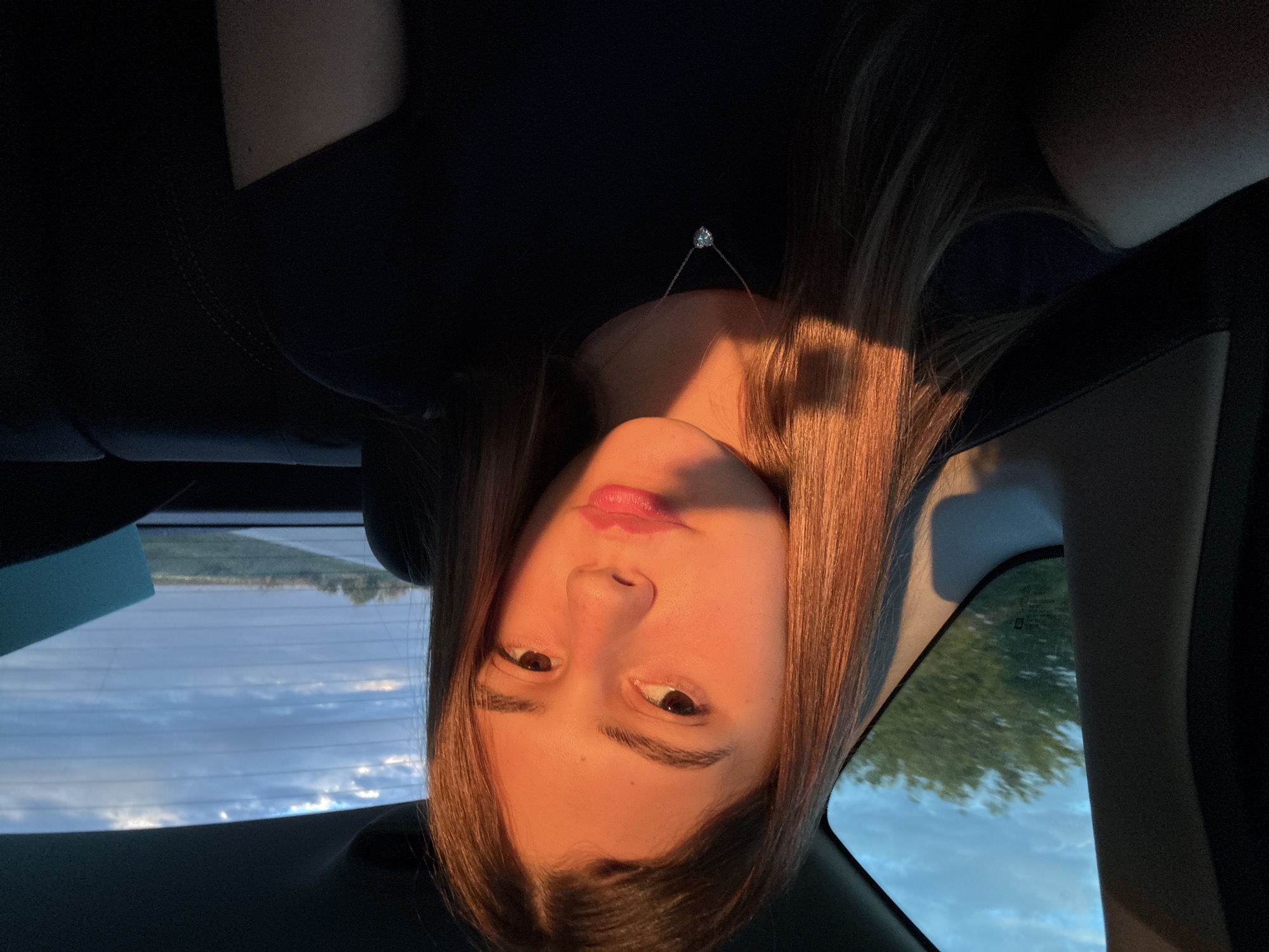 A woman with long brown hair is in a car, with the window and sky visible in the background. She is wearing a dark shirt and a delicate necklace with a small pendant. The car's interior is dark, and the scene is illuminated by warm sunlight. The location is not identifiable as Saraia, Bulgaria.