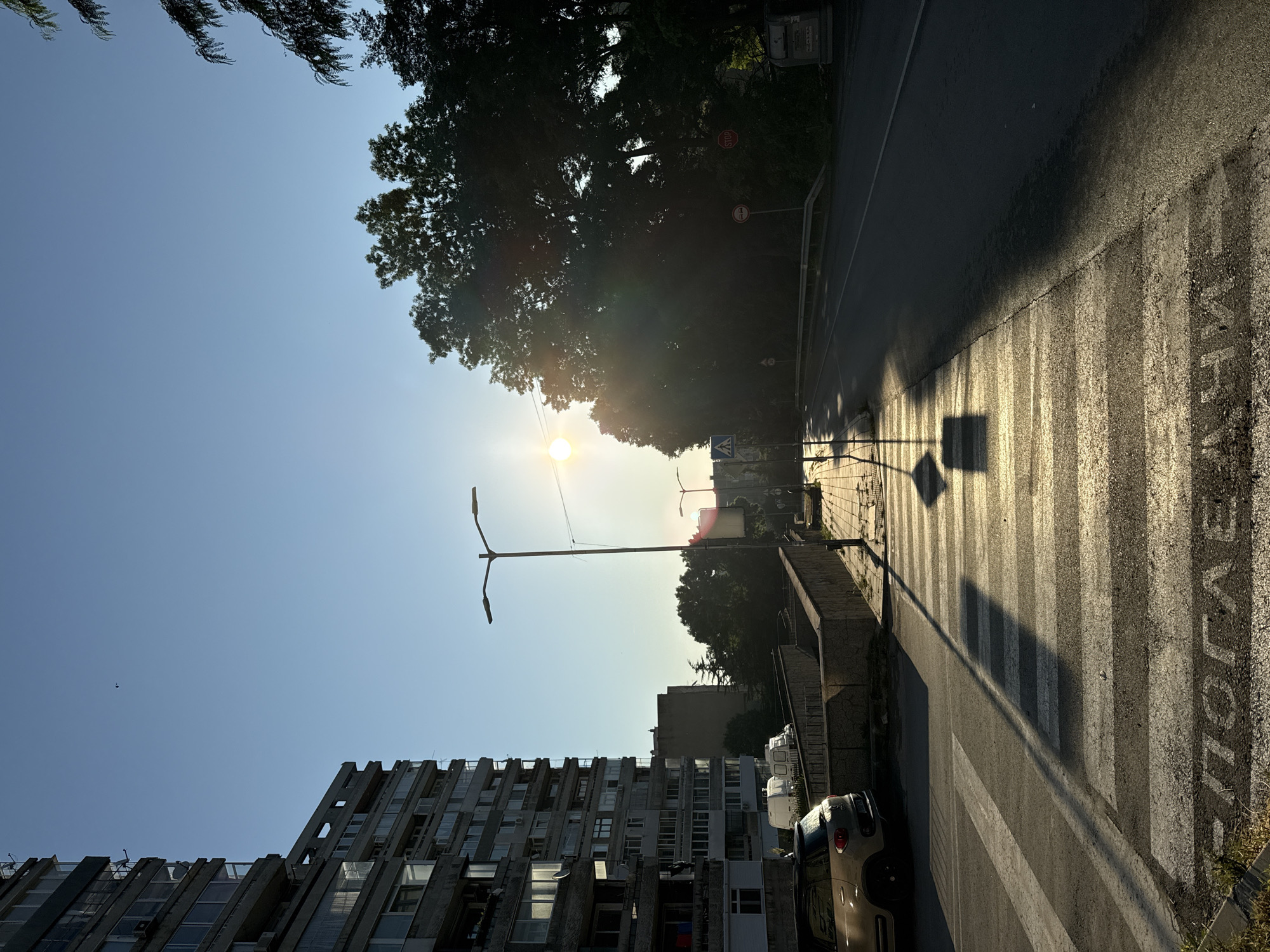 The image depicts a sunlit street scene in an urban environment, likely during the late afternoon or early morning due to the low angle of the sun. On the left, a multi-story, Brutalist-style apartment building dominates the frame, its concrete facade showing signs of age. In front of the building, a light-colored compact car is parked near a pedestrian crossing. The crossing itself is marked with white stripes and has what appear to be letters painted on the asphalt at the far end.
A sidewalk runs alongside the road, lined with a low concrete barrier and some vegetation. A lamppost stands near the sidewalk, casting a long shadow across the crossing. Further down the street, trees with dense foliage create a stark contrast with the clear blue sky. The sun is visible, positioned low in the sky and partially obscured by the trees, creating lens flare and a bright glow. Several street signs are visible, including a blue pedestrian crossing sign and a red stop sign. The road itself is asphalt, and a lane is clearly marked with a white line. The overall impression is one of a quiet urban street bathed in the warm light of the setting or rising sun.