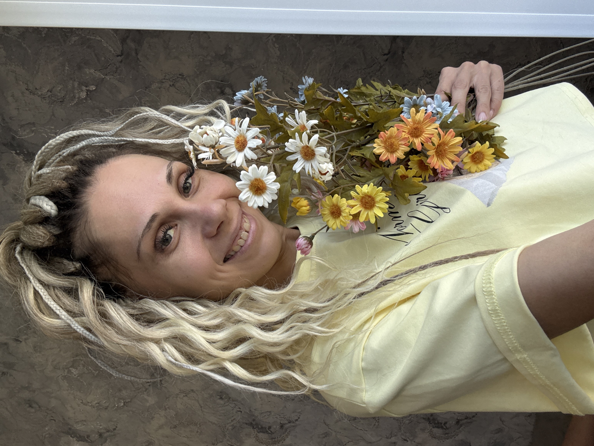 A woman with blonde dreadlocks is lying down against a dark mottled surface, holding a bouquet of mixed flowers (white daisies, orange daisies, yellow daisies, and small blue flowers). She is wearing a pale yellow t-shirt. Her hand is visible at the top right of the frame, holding the flower stems.