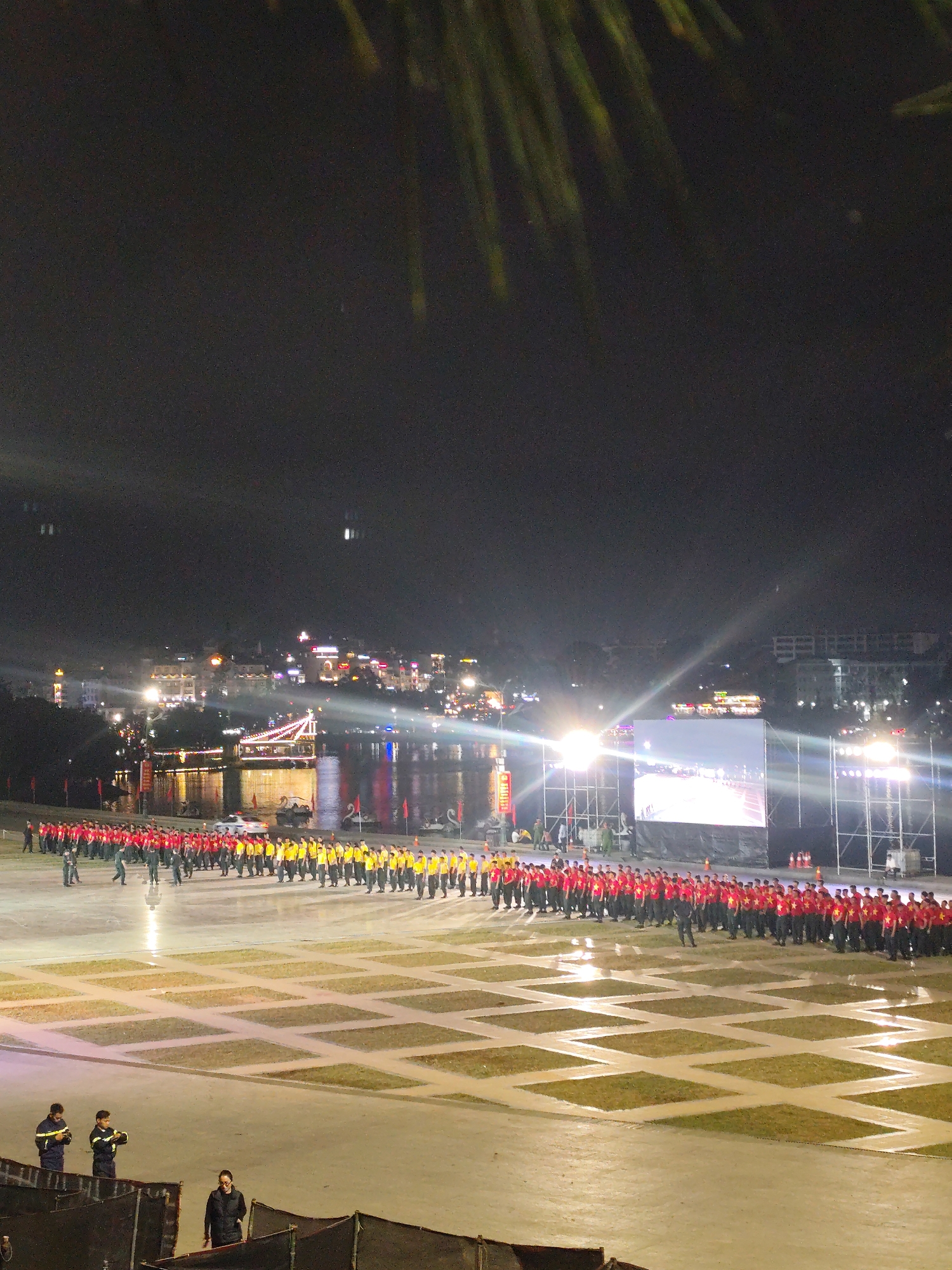 The image depicts a nighttime outdoor event in Da Lat, Vietnam, likely at a public square or plaza near Xuan Huong Lake. A large crowd of people, predominantly young males, are arranged in long, organized rows. Some are wearing red shirts with yellow stars and black pants, while others are in yellow shirts and black pants. They appear to be participating in some form of performance or ceremony.
In the background, the city of Da Lat is visible, illuminated by numerous lights. A prominent feature is a body of water, likely Xuan Huong Lake, with a brightly lit bridge or structure crossing it. Another illuminated structure, possibly a stage or screen, is set up in the middle ground. Several individuals in dark uniforms, possibly security personnel or organizers, are scattered throughout the scene, observing and managing the event. The ground in the foreground is paved with a pattern of squares and illuminated by spotlights. In the lower portion of the image, three individuals in dark work uniforms stand on the paved area. The overall atmosphere is festive and organized, with a sense of anticipation for the event.