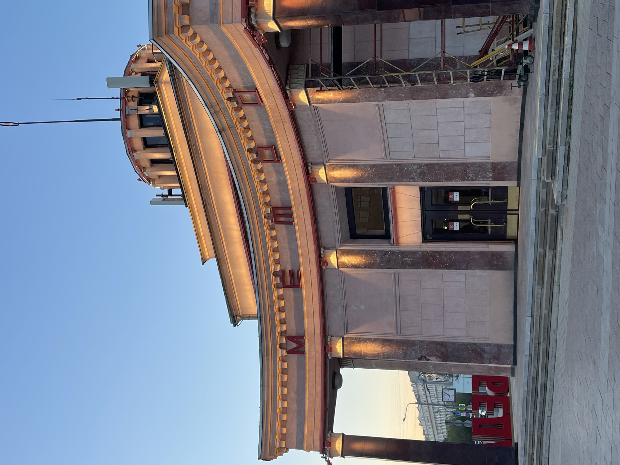 The image captures a section of the Central Armed Forces Museum in Moscow, Russia. The building exhibits a monumental architectural style with a curved facade. The facade is adorned with the Russian word "МУЗЕЙ" (Museum) in large, red, stylized letters. The architecture incorporates decorative columns, textured walls, and a rounded tower-like structure at the top. The image is taken at ground level, looking upwards at an oblique angle. The sky is visible as a clear gradient of blue. The photograph is oriented vertically.