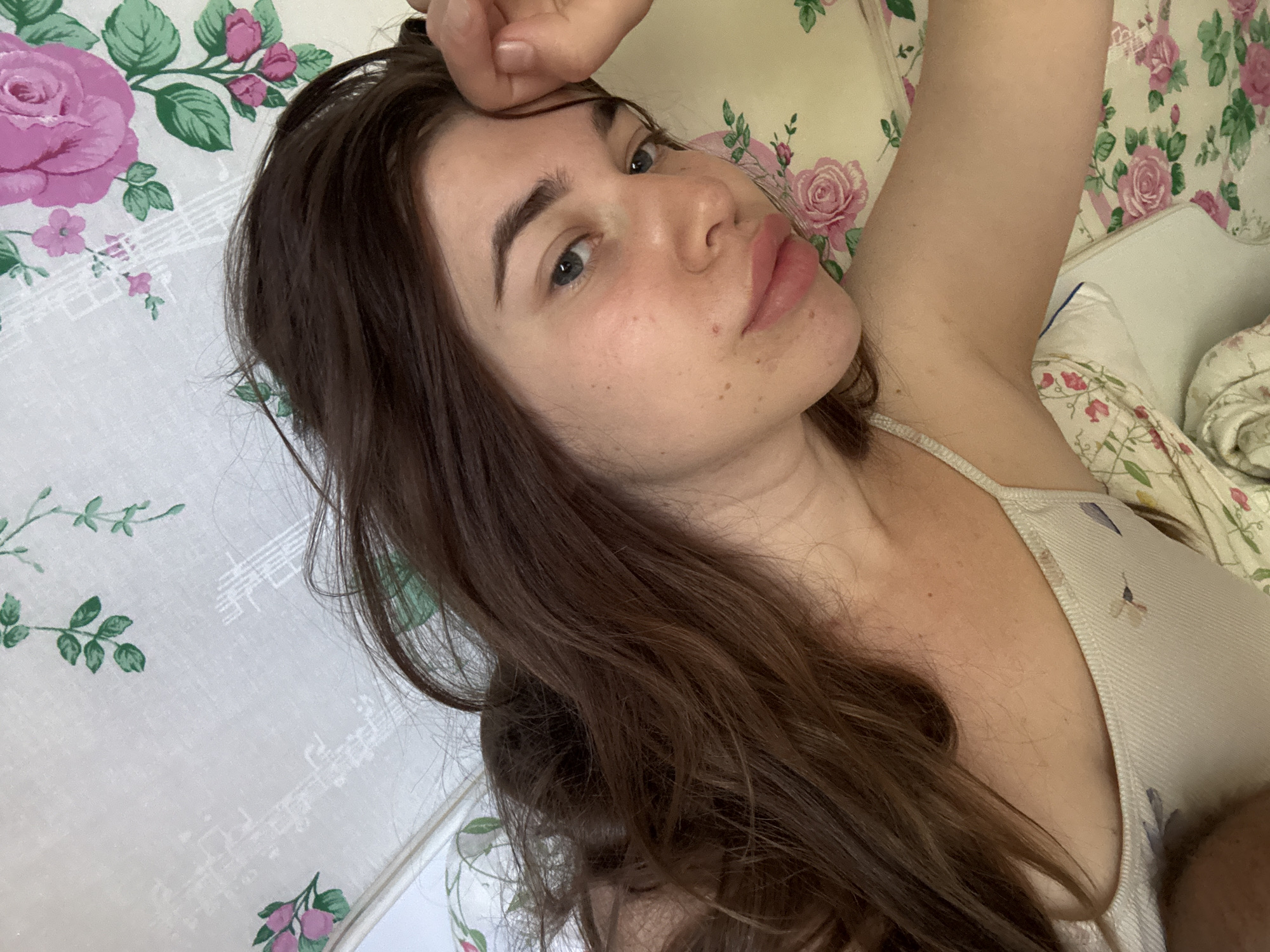 In this close-up, overhead shot, a young woman with long, dark brown hair and fair skin is lying down. She is wearing a light-colored, ribbed tank top with a subtle floral pattern. Her left arm is raised, with her hand resting on her forehead. She has light blue eyes and is looking towards the camera. Her lips are slightly parted and appear to have a pink tint. The background consists of a white fabric with a pattern of pink roses and green leaves, suggesting a floral print bedding or upholstery. The image appears to be taken indoors, possibly in a bedroom. The location of Мещёры, Russia, is not discernible from the visual information provided in the image.