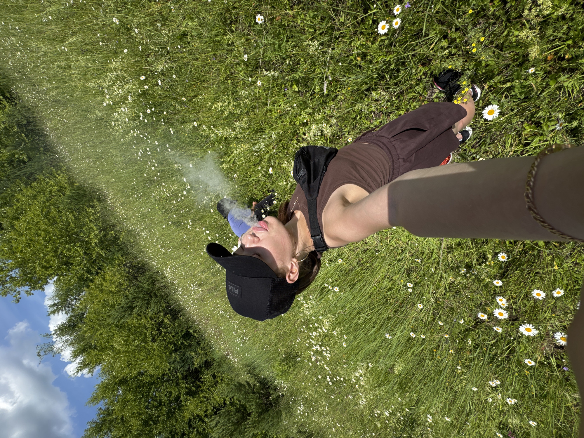 A young woman wearing a black baseball cap, brown tank top, and brown shorts is lying on her back in a field of green grass and white wildflowers. She is holding up her right arm, obscuring part of the image, and is exhaling a puff of smoke. A black fanny pack is worn across her chest. The background consists of lush green trees and a blue sky with white clouds. The location is identified as Garevaya, Russia.