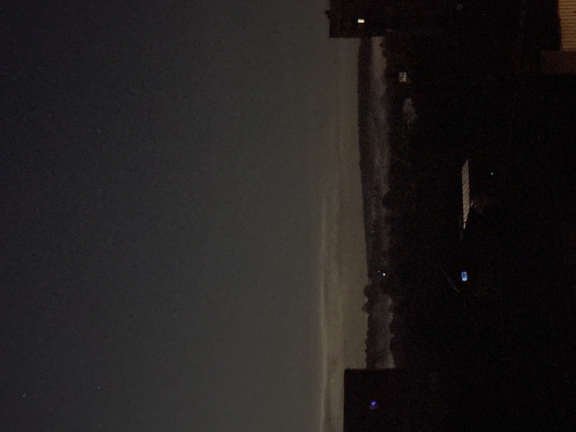 The image shows a nighttime view of what appears to be a city skyline. The left side of the image is predominantly dark grey, possibly the night sky. On the right side, silhouetted buildings and trees are visible, obscuring a portion of the sky. Some lights, both yellow and blue, are visible in the buildings. The location is undetermined, as the city is unknown.
