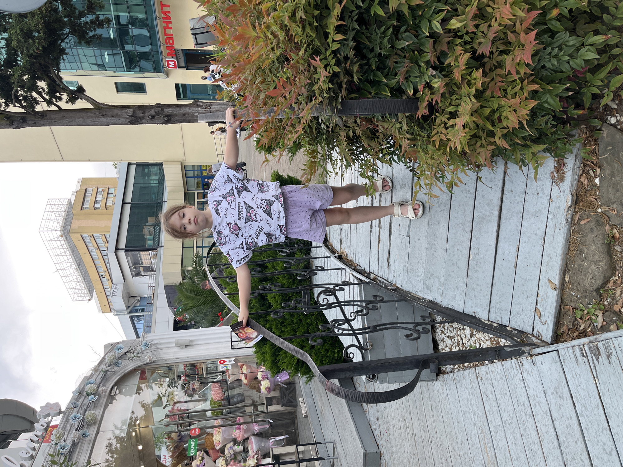 A young Caucasian girl with light brown hair stands on a wooden bridge with a weathered, gray painted surface. She is facing forward, with her arms outstretched to her sides, and her left hand is touching the bridge railing. The girl is wearing a white, cropped t-shirt with a pattern of cartoon characters and light purple shorts with a polka dot pattern. She has white sandals on her feet.
To the left of the girl, there is a white archway adorned with artificial blue flowers and greenery, leading to a storefront with a large glass window displaying an arrangement of bouquets. A sign in Russian is visible through the window, with the word "ОТКРЫТО" (Open) in green.
To the right of the girl, a dense green bush with red and orange-tipped leaves occupies the foreground. The bridge railing is made of black wrought iron with decorative scrollwork.
In the background, several multi-story buildings are visible. One building on the right has a large red and white sign that reads "М МАГНИТ" in Russian, indicating a store. The sky is overcast. The scene appears to be an outdoor public space, possibly a commercial or pedestrian area in Sochi, Russia, given the signage.