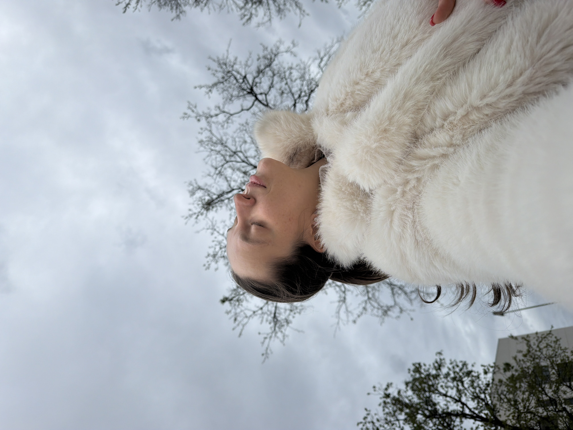 A woman with closed eyes, wearing a white furry coat, is facing upwards. The background shows a cloudy sky and bare tree branches. A building is partially visible in the lower right corner. The location cannot be definitively determined to be Budva, Montenegro from this image alone.