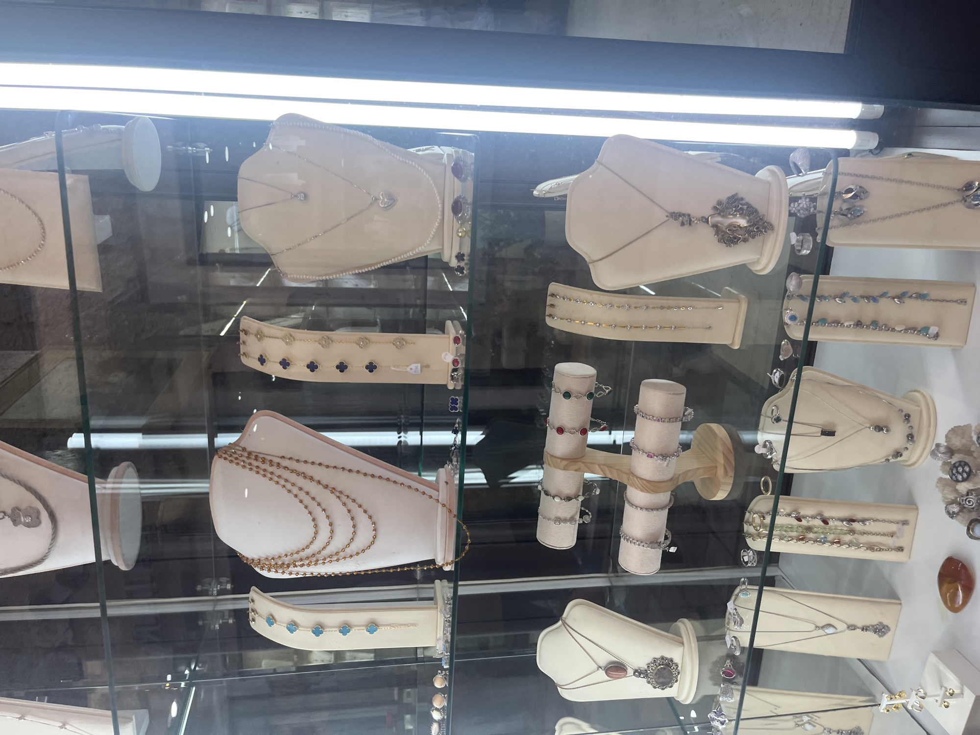 This is a media file showing a display of jewelry inside a store. The jewelry, consisting of necklaces and bracelets, is presented on white mannequins and stands. The items appear to be made of various metals and precious stones, including diamonds, pearls, and colorful gems. The lighting is bright, suggesting artificial light, possibly fluorescent tubes visible at the top of the frame. The setting is a retail environment, likely a jewelry store, with glass shelves and reflections indicating a display case. The time of day cannot be determined from the image. There is no visible text. The context is Budva, Montenegro.