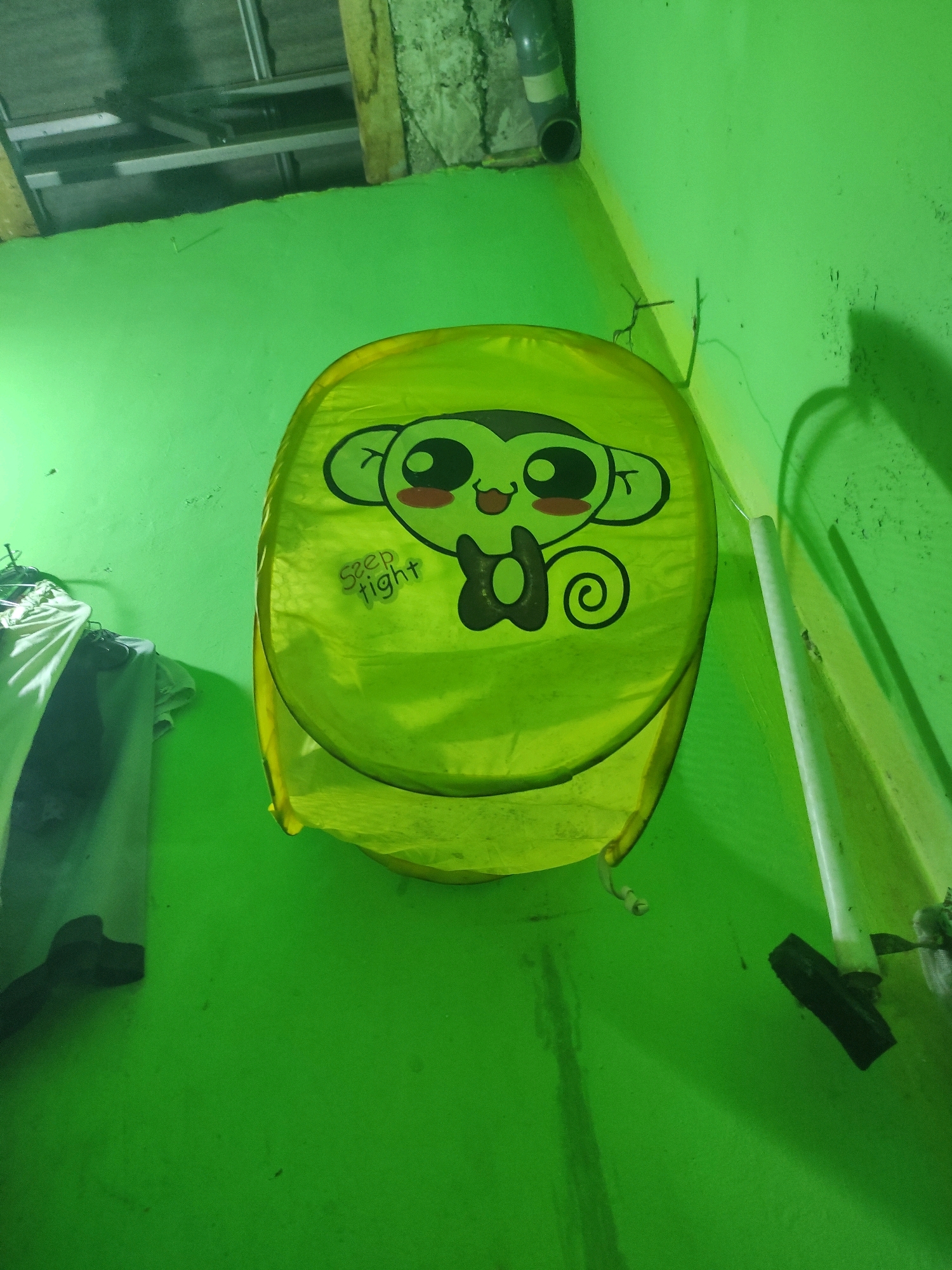 The image shows a lime green laundry basket with a cartoon monkey face printed on it. The monkey has large eyes, rosy cheeks, and a smiling mouth. The text "sleep tight" is also visible on the basket. The basket is standing upright against a bright green wall, which appears to be painted. In the background, a white pipe is visible attached to the wall. To the left of the basket, there are some clothes hanging on a rack. The lighting in the scene is dim and casts shadows, suggesting it might be nighttime or an indoor location with limited light. The environment appears to be a somewhat cluttered storage area, possibly a laundry room or closet, in a residential setting. Without further information, it is not possible to confirm the location as Padalarang, Indonesia.