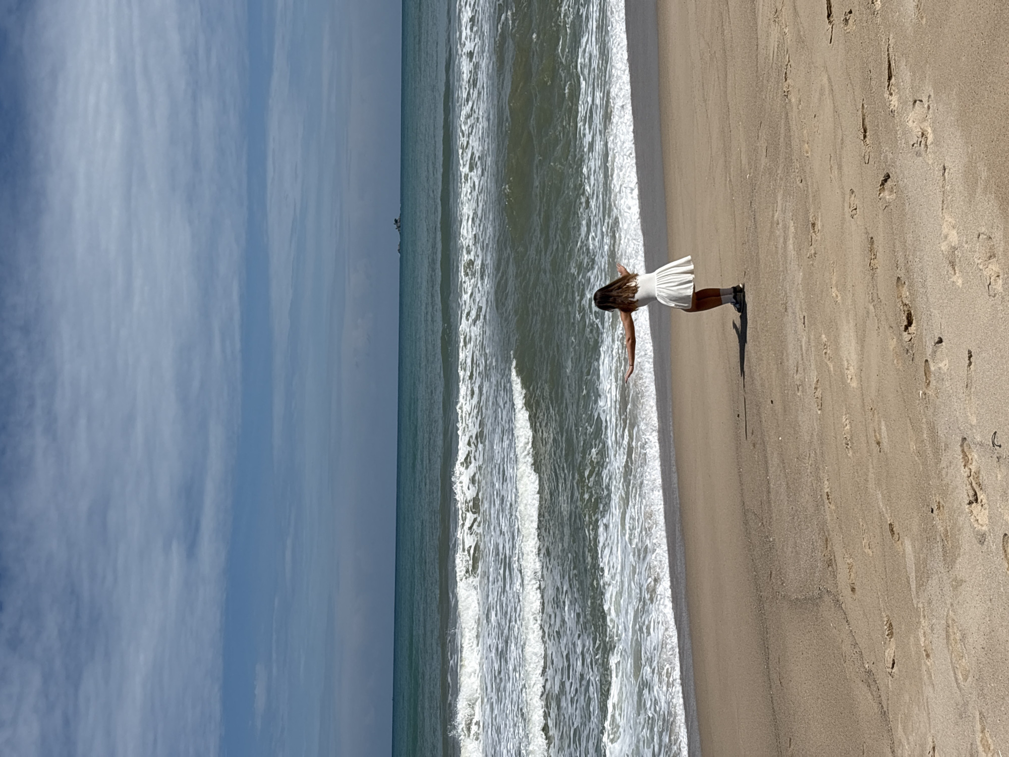 A person wearing a white dress stands on a sandy beach with their arms outstretched. The ocean, with rolling waves, is in the background. The sky is blue with scattered clouds. A small island or rock formation is visible in the distance on the horizon. The scene appears to be located in Choeng Thale, Thailand.