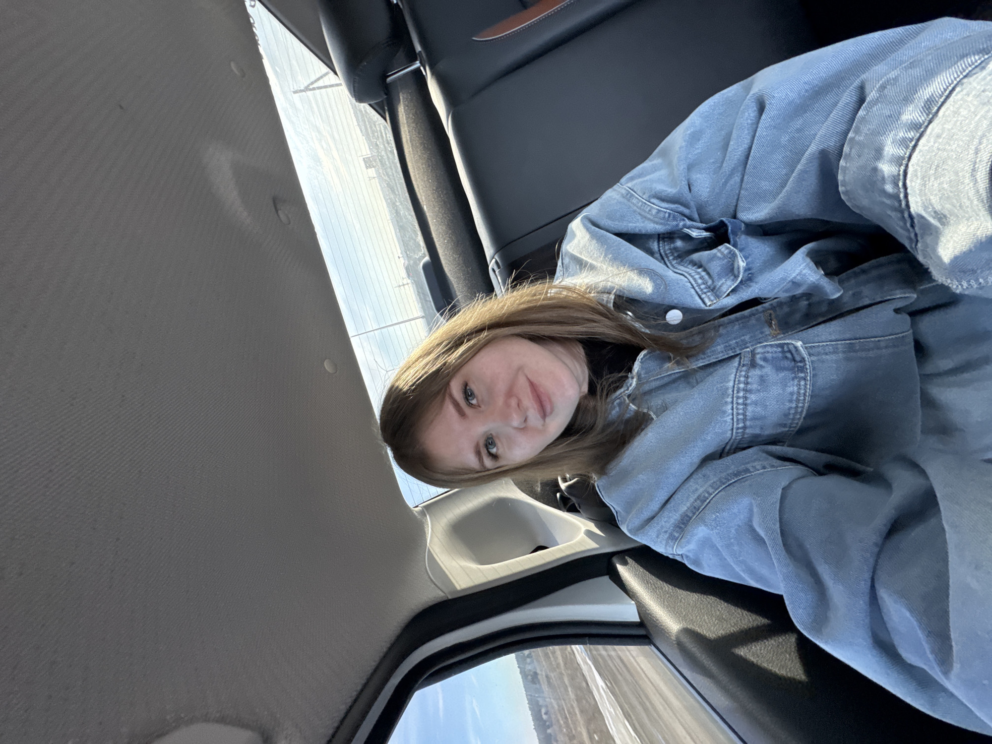 A young woman sits inside a vehicle, possibly a car, while driving through the outskirts of what is probably Perm, Russia. She wears a denim jacket and appears to be looking at the camera, maybe taking a selfie. The environment outside is visible through the car windows, including a barren landscape under a partly cloudy sky. The interior of the vehicle is modern, with dark leather seats and light-colored ceiling.