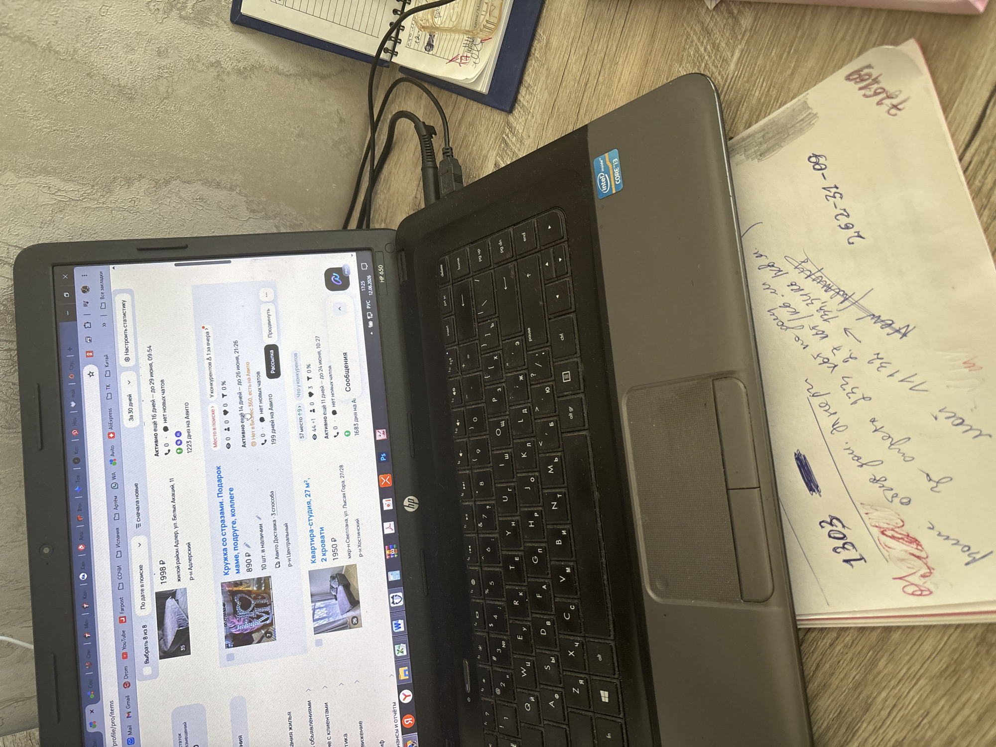 The image shows a laptop open on a wooden desk. On the laptop screen, an online marketplace interface is visible, displaying various items for sale, including furniture, a decorative item, and an apartment. The location of Sochi, Russia is mentioned in one of the item descriptions: "жилой район Адлер, ул. Белых Акаций, 11 р-н Адлерский". A handwritten note is placed on the desk in front of the laptop, with what appear to be numbers and some scribbled text. The overall environment appears to be a home office or living space.