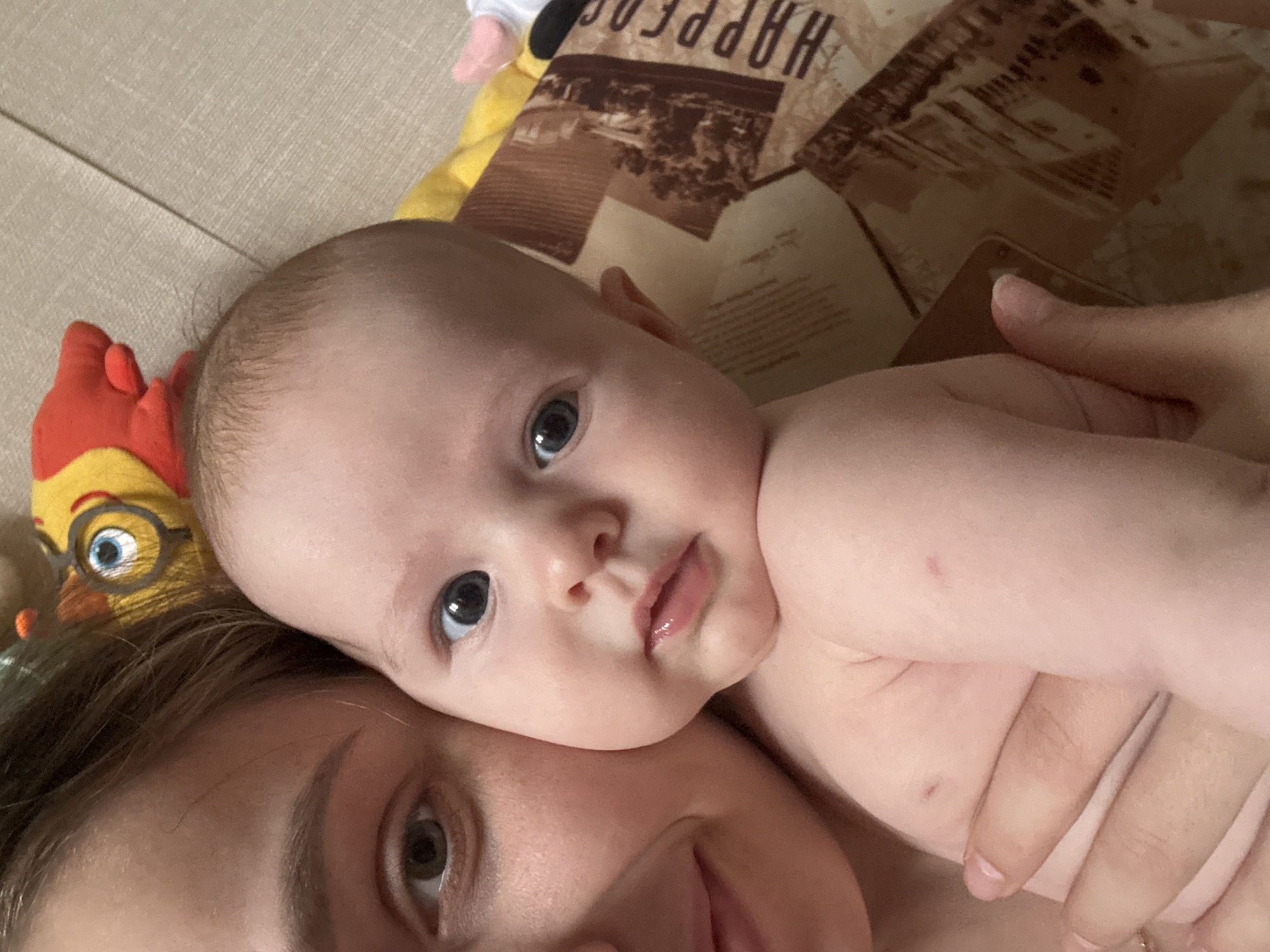 The media file captures an intimate, close-up moment between an adult and a baby, likely taken indoors in a home setting. The image is oriented as if viewed from above, with the subjects lying down.
The central subject is a baby, visible from the chest upwards, with a prominent focus on its face. The baby has fair skin, sparse light hair, and striking blue eyes that gaze directly at the camera. Its lips are slightly parted, giving a serene expression. A small, red and yellow plush toy, resembling a cartoon chicken or bird with large blue-irised eyes and painted eyeglasses, is situated next to the baby's head.
A portion of an adult's head and face, with brown hair and brown eyes, is visible in the lower left section of the frame, positioned intimately close to the baby. An adult hand is seen gently cradling the baby, suggesting a tender embrace or cuddle. One of the detailed crops shows a small red mark or scratch on the baby's delicate skin, possibly on its hand or forearm.
The background consists of a patterned blanket or fabric featuring vintage-style prints, resembling old photographs or postcards, and various texts. The word "HAPPY" is clearly legible in uppercase, stylized lettering on this fabric. The lighting appears soft and natural, suggesting a daytime scene. The overall atmosphere conveys warmth, affection, and peaceful companionship between the adult and the infant in Chaikovsky, Russia.