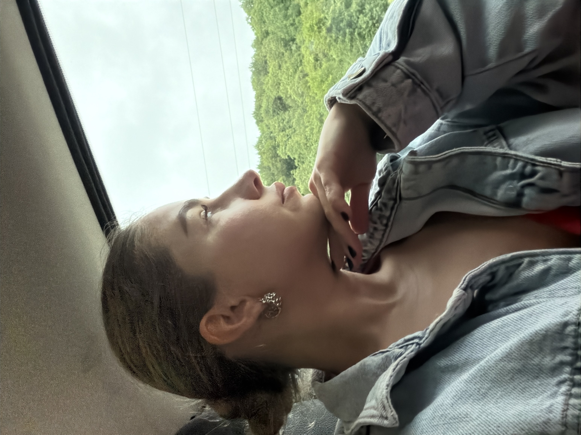A young woman with dark hair pulled back is seen from the side, looking out a window. She is wearing a light blue denim jacket over a red top and has a silver earring in her left ear. Her right hand rests on her chin. Outside the window, a dense green forest is visible under an overcast sky. The interior of what appears to be a vehicle is visible in the foreground, with a textured ceiling and a dark seat. The location is not specified to be in Gornji Vakuf-Uskoplje, Bosnia and Herzegovina.