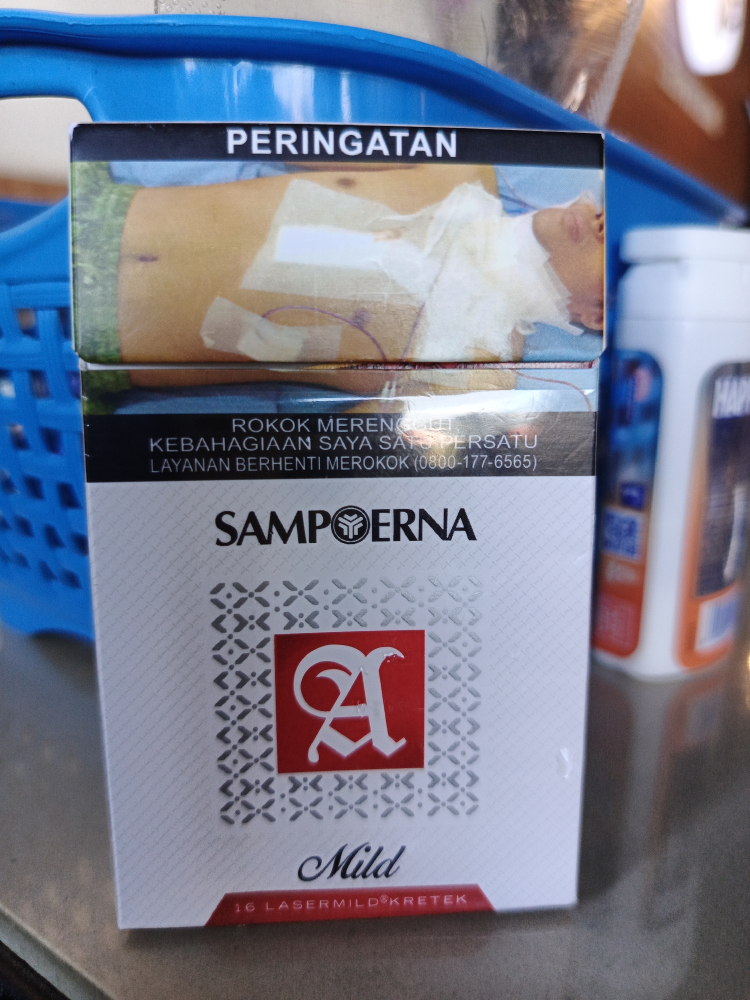 A close-up, eye-level shot shows a pack of Sampoerna A Mild cigarettes positioned vertically in the foreground, resting on a brown textured surface. The cigarette pack is white with a patterned design and features the brand name "SAMPPOERNA" prominently. A stylized red letter "A" is visible within a red square in the center of the pack. Above the brand name, a black band contains text in Indonesian, including "PERINGATAN" (Warning) and information about quitting smoking. The upper portion of the pack displays a graphic image of a person's torso with medical bandages.
Behind the cigarette pack, a bright blue plastic basket is visible on the left, and a white bottle with an orange label (partially obscured) is on the right. The background is blurred and out of focus, suggesting an indoor or shaded outdoor environment. The scene appears to be a still life arrangement, with no people or discernible action taking place. The lighting is adequate, highlighting the details of the cigarette pack. The overall impression is that of a product shot, possibly taken in a market or shop setting. The presence of a blue basket and a white bottle suggests a retail or domestic context, potentially in Bireuenmeunasahdayah, Indonesia, given the product's origin.
