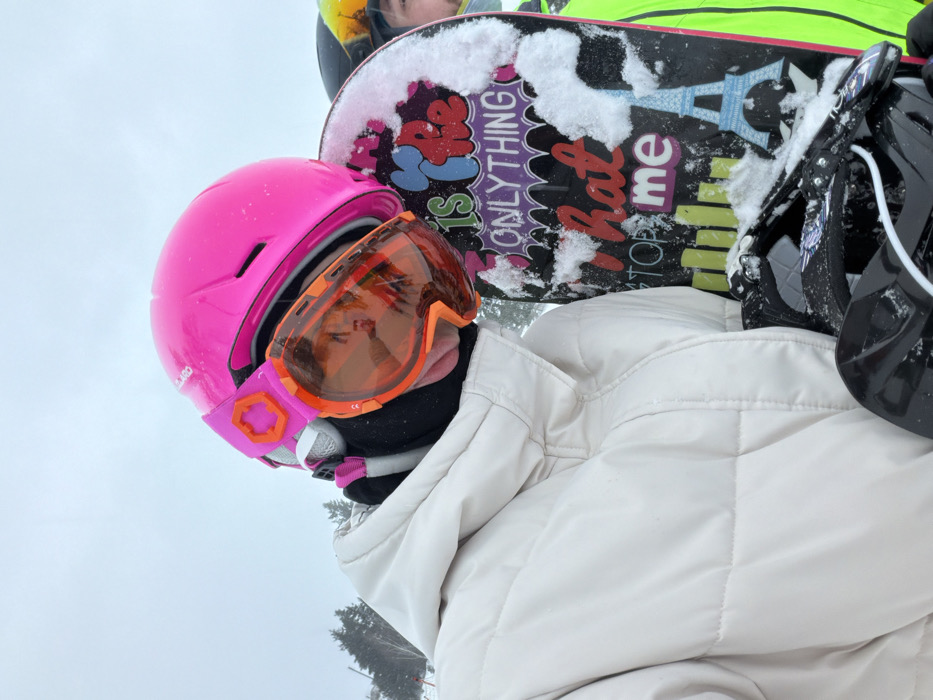 A person is wearing a pink helmet with orange ski goggles. They also have a white puffy jacket and a black neck gaiter. A snowboard with a black background and colorful text is visible behind them. The snowboard has snow on it, and the text includes phrases like "Is ONLYTHING" and "hate me". There is another person with a yellow helmet and a second helmet with a black color and a pair of snowboards in the background.