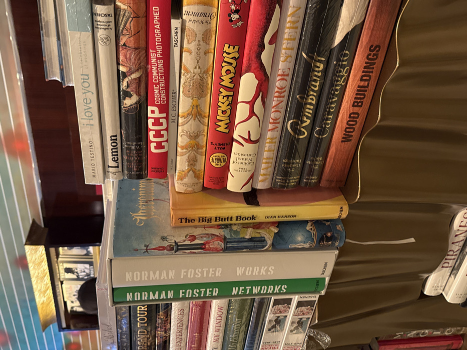 The image displays a shelf laden with books. Visible titles include "I love you" by Mario Testino, "Lemon," "CCCP" (Cosmic Communist Constructions Photographed), M.C. Escher's works, "Mickey Mouse," "Mailer Monroe Stern," "Rembrandt," "Caravaggio," "Wood Buildings," "The Big Butt Book" by Dian Hanson, "Norman Foster Works," and "Norman Foster Networks." Other books are partially visible with titles like "Grand Tour" and "My Education." The spines exhibit a range of colors and typography, suggesting a diverse collection of art, photography, and architecture publications.