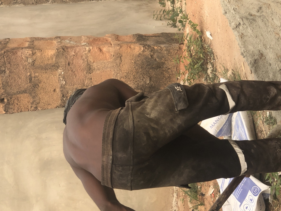 An individual, observed from the back, is engaged in manual labor outdoors in Uke, Nigeria. The person has dark skin, is shirtless, and wears heavily soiled, dark-colored cargo pants with a visible "AE" logo on a side pocket and lighter reflective or worn patches near the knees. A dark cloth is tied around their head. The individual is bent forward, implying an action such as shoveling or mixing, with a tool handle visible near their left leg. Behind the person, two white sacks, identified as cement bags with partial text including "MANGAN," "CPM," and "CEK," rest on the reddish-brown earth. In the foreground, a pile of light-grey material, consistent with sand or concrete mix, is present. The background features a light-colored, possibly newly plastered wall section on the left, and a deteriorating reddish-brown brick pillar or wall section on the right. Sparse green vegetation and scattered debris are visible on the ground. The scene depicts an active construction or renovation site.