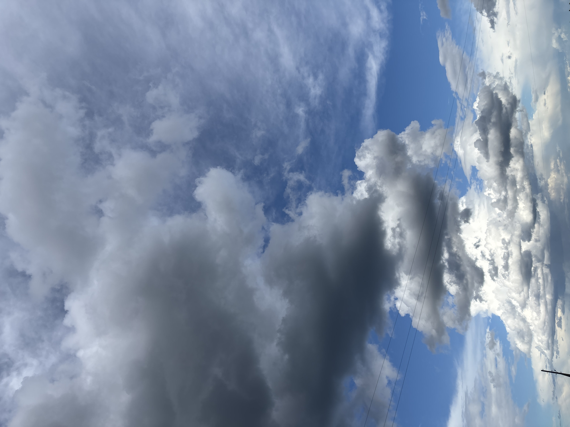 A dynamic sky over the city of Kurgan, Russia is depicted in this image. Large, dark cumulus clouds dominate the left side of the frame, hinting at impending weather. To the right, lighter, wispy clouds streak across a vibrant blue sky, suggesting a mix of atmospheric conditions. Power lines stretch horizontally across the scene, subtly indicating the presence of urban infrastructure. The bottom portion of the image shows a glimpse of the horizon, with faint outlines of what could be distant structures or natural formations. The overall impression is one of natural beauty and the vastness of the sky.