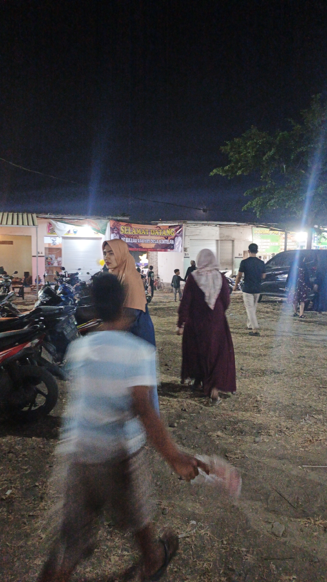 The image captures a nighttime scene at a market in Ketemasdungus, Indonesia. A banner reading "SELAMAT DATANG" (Welcome) is visible in the background, indicating the entrance to a "BAZAR RAKYAT DESA KINTELAN" (People's Market of Kintelan Village). Several people are present, including a woman wearing a light brown hijab and a dark dress, standing near a row of motorcycles. Another woman in a dark maroon abaya and a light-colored hijab walks away from the camera. A man in a black t-shirt and light-colored pants walks towards a dark-colored vehicle. In the foreground, a person wearing a light blue and white striped t-shirt and shorts is blurred due to motion, walking away from the camera and holding a translucent plastic bag. The ground is covered with dry grass and dirt, and a tree with green foliage is visible on the right side of the image. Lights illuminate the area, suggesting the market is open at night.