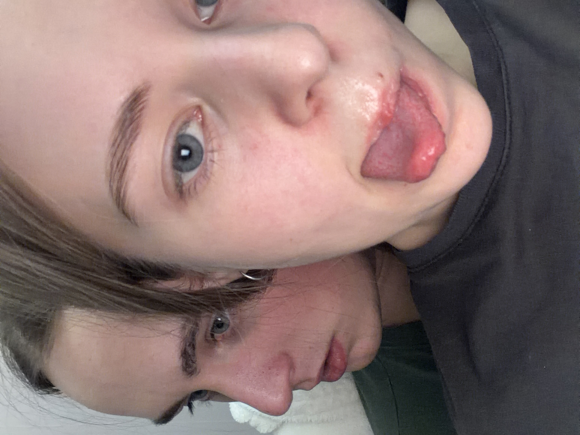 This close-up portrait features two young individuals, likely taken indoors in Perm, Russia. The image is oriented diagonally, with the head of one person stacked partially above the other.
The person at the top has fair skin, light blue eyes, and light brown hair and eyebrows. Their face is angled towards the camera, and they are playfully sticking their tongue out, which partially covers their upper lip and the area below their nose. There appears to be some redness around their mouth/nose area where the tongue meets the skin. They are wearing a dark grey or black top, visible on the right side of the frame. Their expression conveys playfulness.
Positioned below and slightly behind the first person is a second individual. They also have fair skin, with blue-grey eyes, and darker brown hair and eyebrows. Their head is tilted to the left, and they have a small silver hoop earring visible in their ear. Their expression is calm and neutral, with slightly parted lips. They are wearing a dark green top, and a fluffy white texture, possibly a blanket or pillow, is visible near their head in the lower-middle part of the image.
The photograph captures a lighthearted, intimate moment between the two, with the top person engaging in a playful gesture. No text is visible in the image.