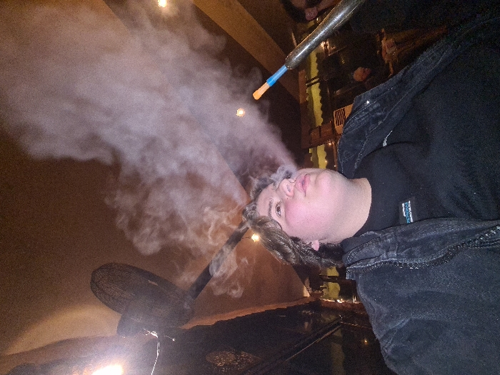 A person exhales a large plume of smoke. The smoke is thick and white, obscuring the background. The person is wearing a black shirt and denim jacket. A hose with a blue and orange tip is visible. The scene appears to be indoors, possibly in a dimly lit hookah lounge or similar establishment.