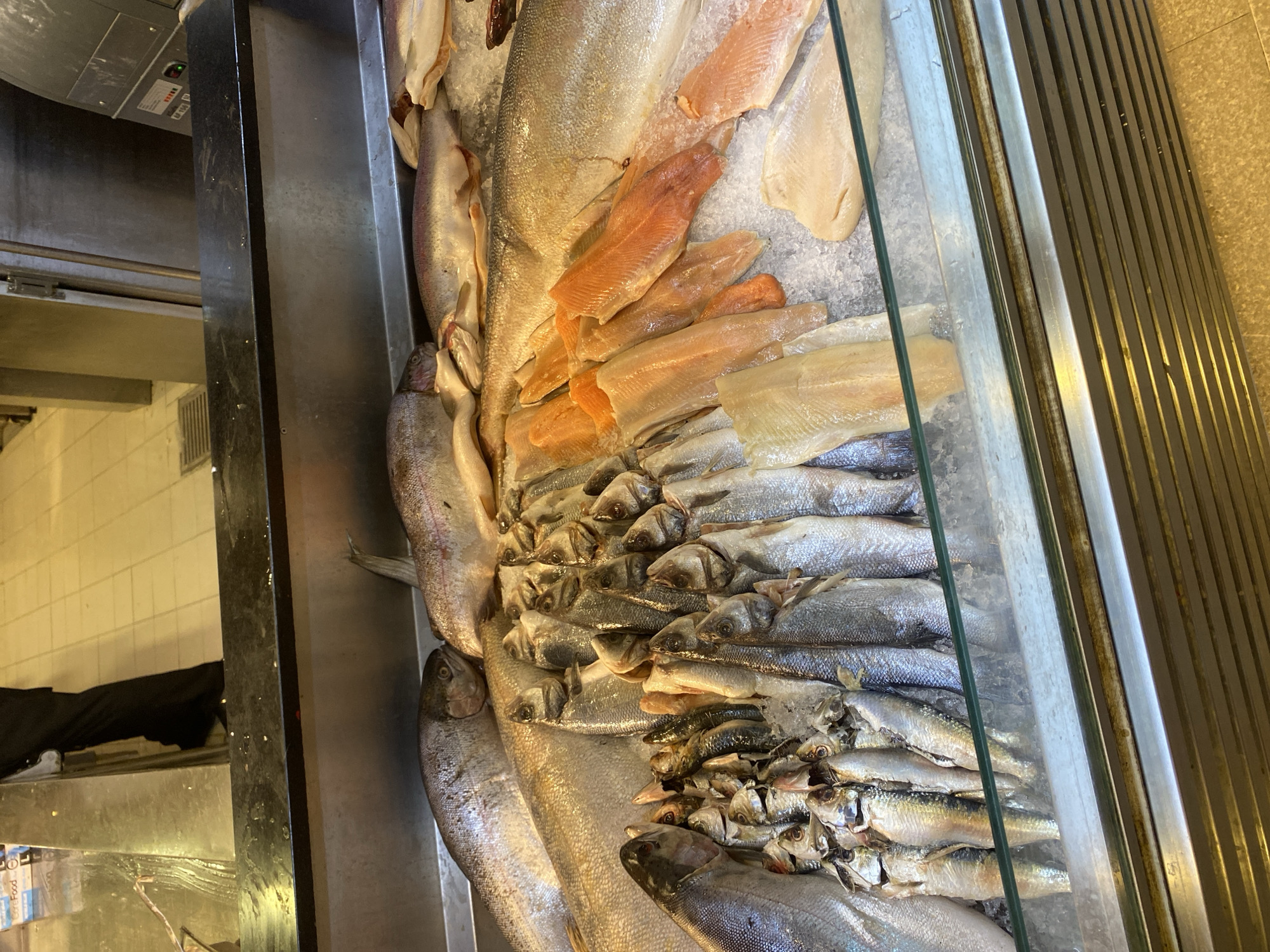 The image shows a display case filled with various types of fresh fish on ice. In the foreground, a large glass barrier separates the viewer from the fish. Several whole fish, including what appear to be salmon or trout due to their size and coloration, are laid out. Smaller silver fish, possibly sardines or anchovies, are piled in front of the larger fish. There are also several rows of fish fillets, some with a reddish-orange hue and others paler, laid out in distinct sections. The fish are arranged on a bed of crushed ice, suggesting they are being prepared for sale in a market or fishmonger. In the background, a person wearing black trousers and a dark top is visible, standing behind the display and appearing to be either a vendor or an employee. The flooring in the background is tiled. The environment suggests a retail setting, possibly a fish market or a seafood counter in a larger store. The city of Aksu, Turkey, is not explicitly identifiable within the image's content.
