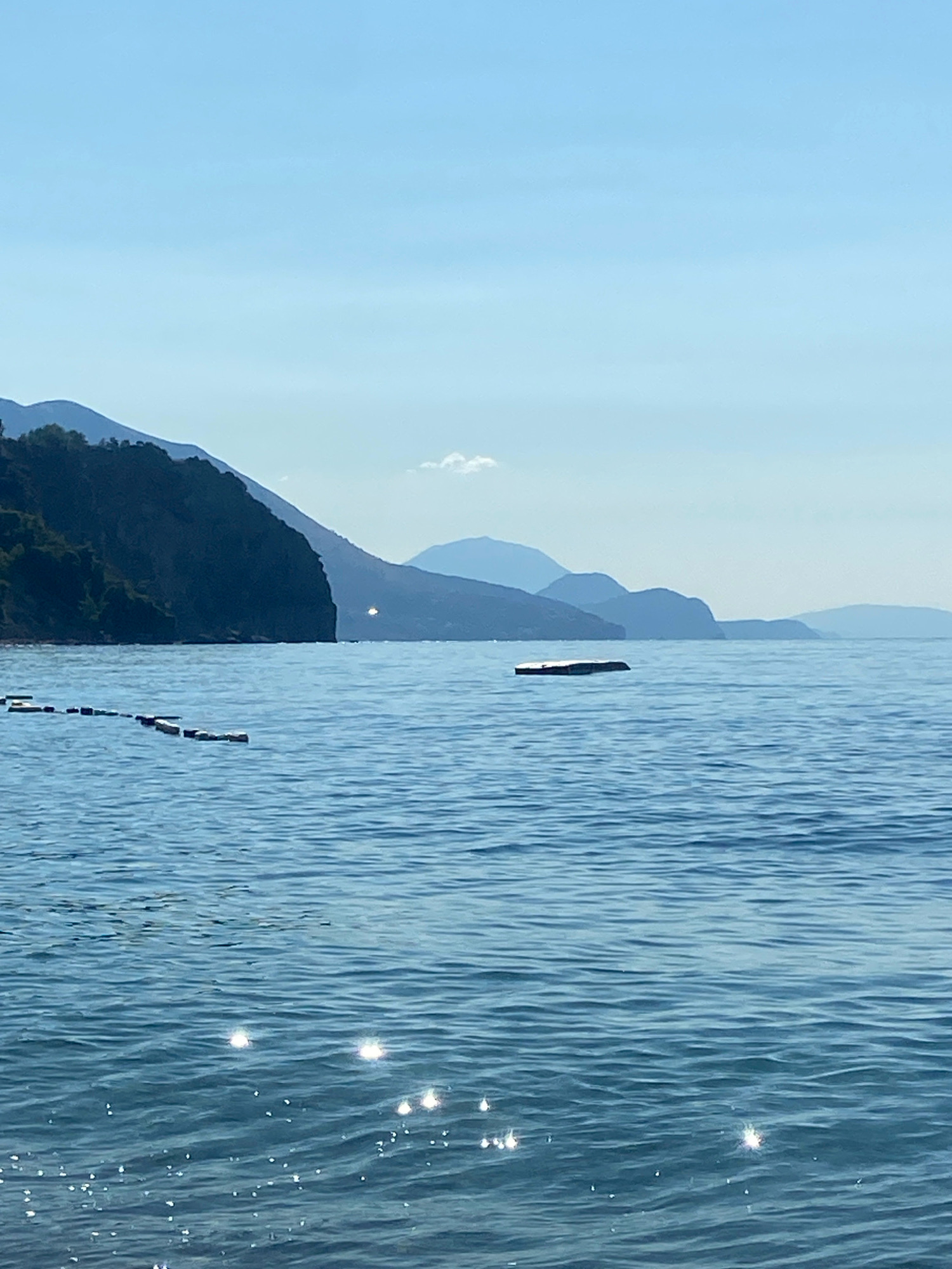 A tranquil, wide shot captures a calm sea under a clear blue sky, with layers of hazy mountains in the background. In the foreground, the water is a deep blue, reflecting the sunlight with numerous bright sparkles. A series of white buoys, connected by a rope, are visible on the left side of the frame, marking a boundary in the water. Further out, a rectangular, light-colored floating object rests on the surface of the sea. The dark, tree-covered mountains on the left are sharply defined, contrasting with the softer, muted tones of the distant mountain ranges. A small, white cloud drifts in the sky above the horizon. The overall impression is one of serene natural beauty, likely a coastal scene near Prijevor, Montenegro.