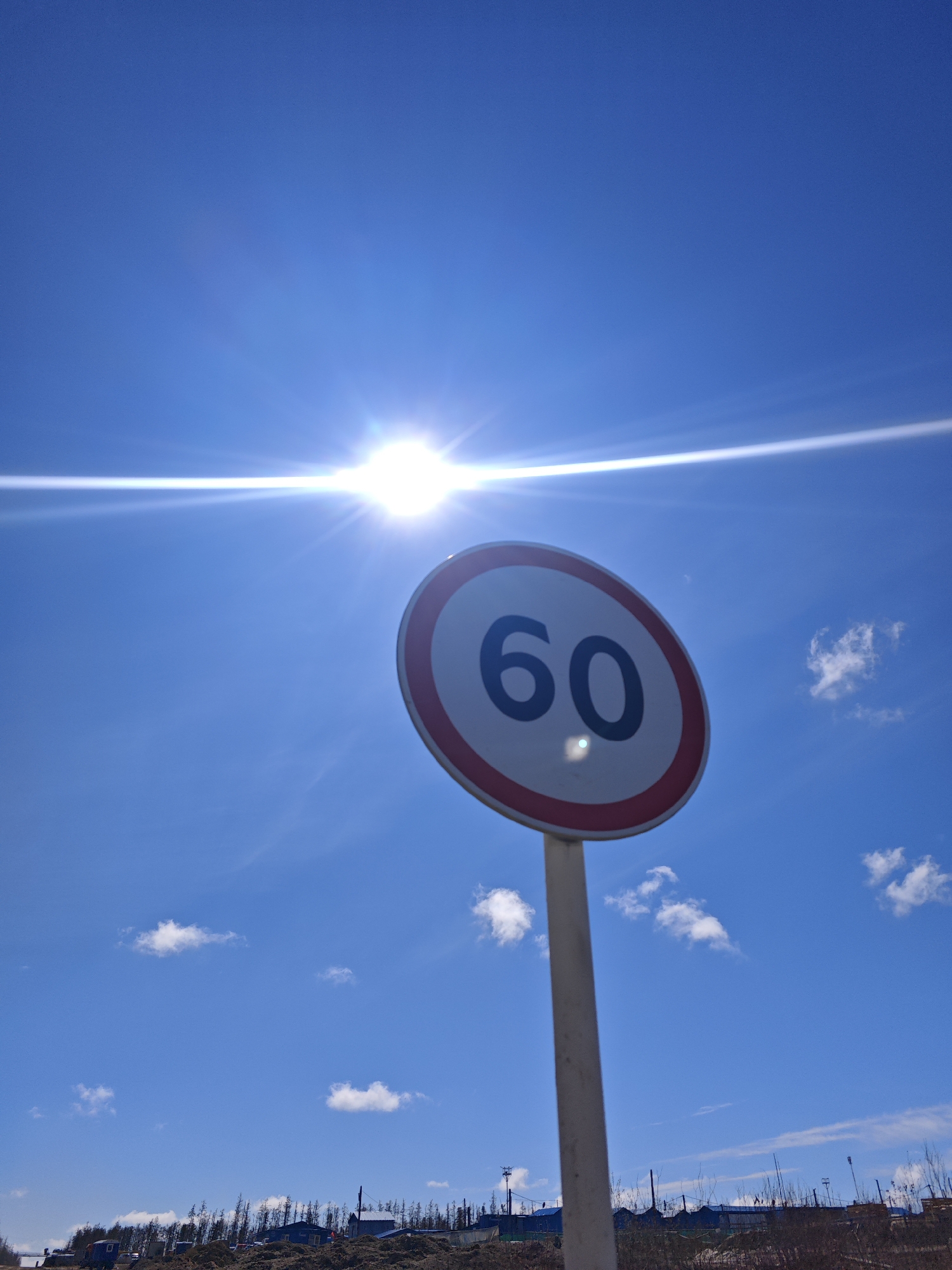 The image shows a speed limit sign of 60 km/h against a bright blue sky with a few scattered clouds. The sun is shining directly above the sign, causing lens flare. In the background, there is a landscape with small buildings and trees, possibly indicating a rural or semi-urban area. The city and region are noted as Unknown, Russia.