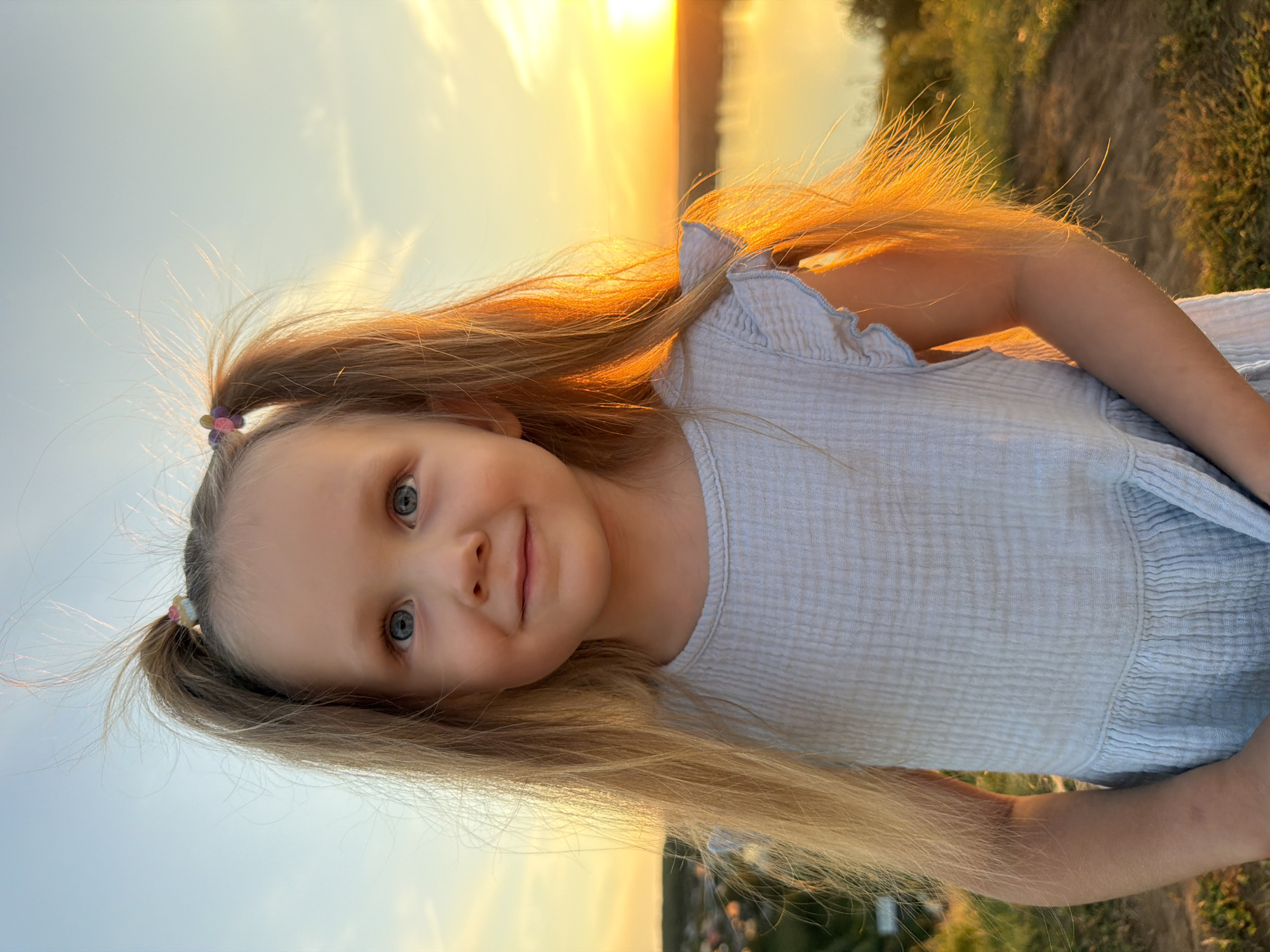 The image shows a close-up, sideways portrait of a young girl with long, blonde hair. She has bright blue eyes and a slight smile. Her hair is tied into two pigtails with small, colorful hair ties. She is wearing a light blue, textured dress with ruffled sleeves. The background is blurred, showing a field of grass and a sunset with warm, golden light. The overall mood is cheerful and peaceful, capturing a moment of simple joy in an outdoor setting. The time of day is sunset, indicated by the golden hour light. The location is suggested to be Olkhovka, Russia. There is no visible text in the image.
